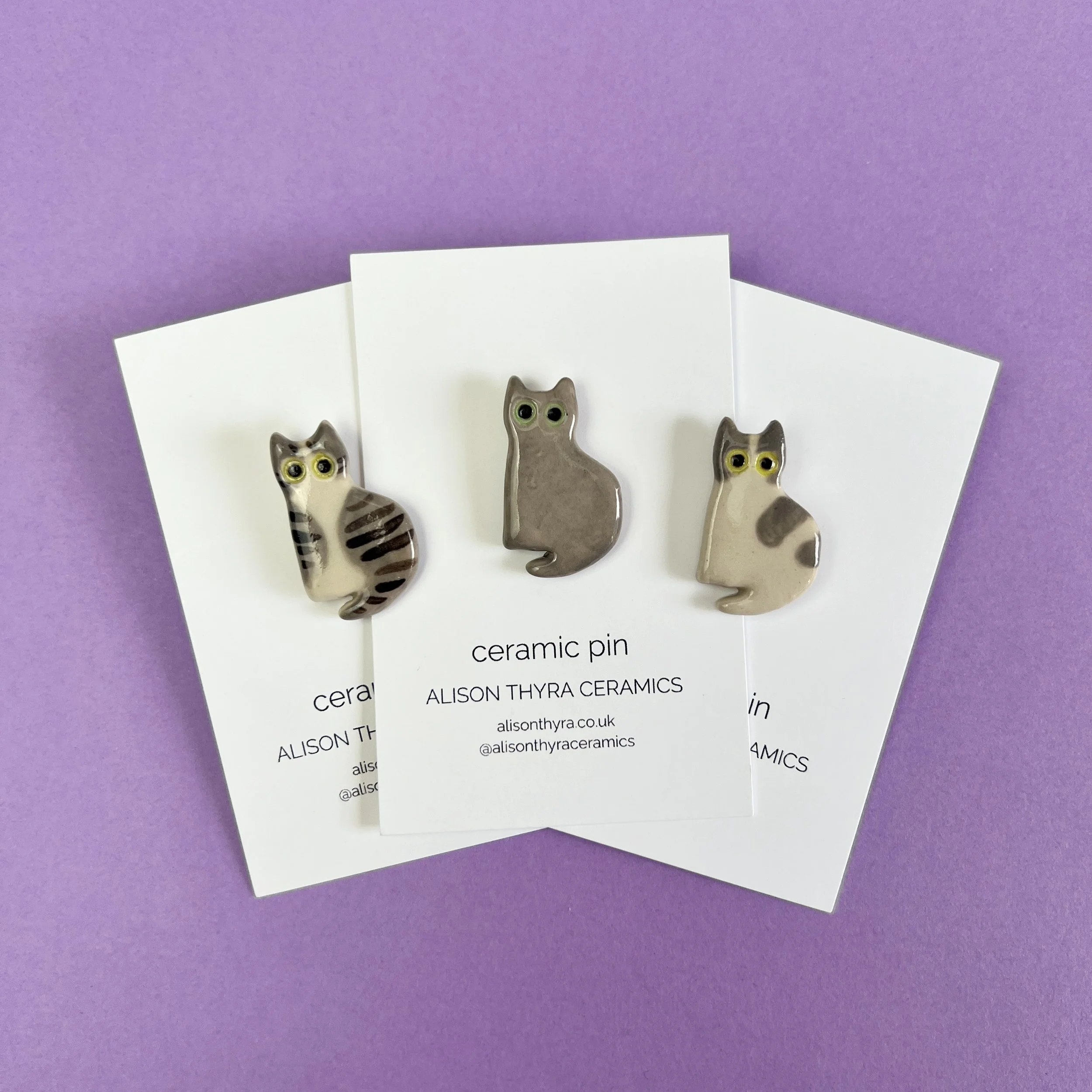 Three ceramic cat pins in different colors and patterns on white Alison Thyra Ceramics backing cards with black text, set against a purple background.
