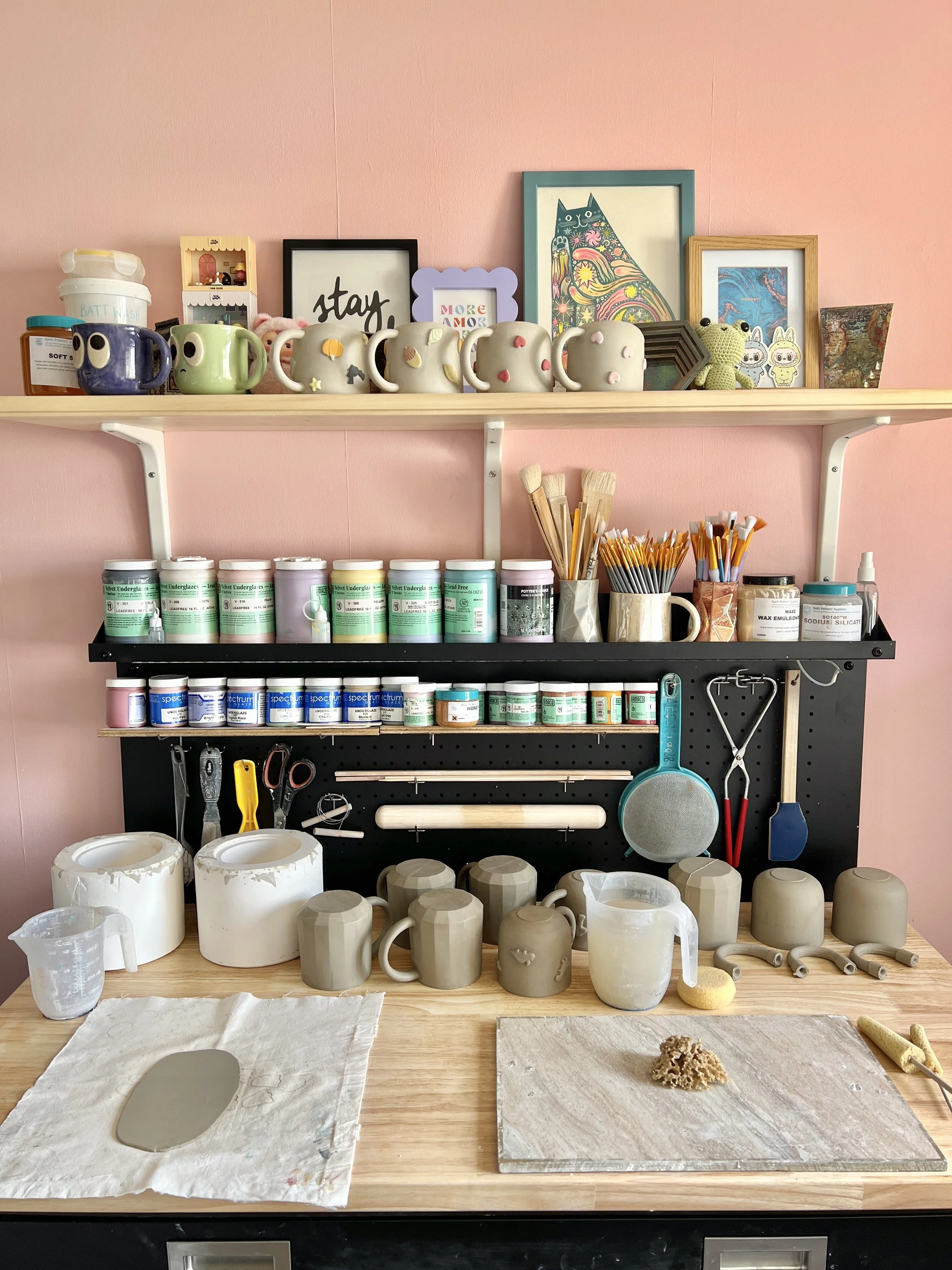 A pottery workspace with shelves holding decorated mugs, art frames, and art supplies, and a work table with unglazed clay mugs and tools.