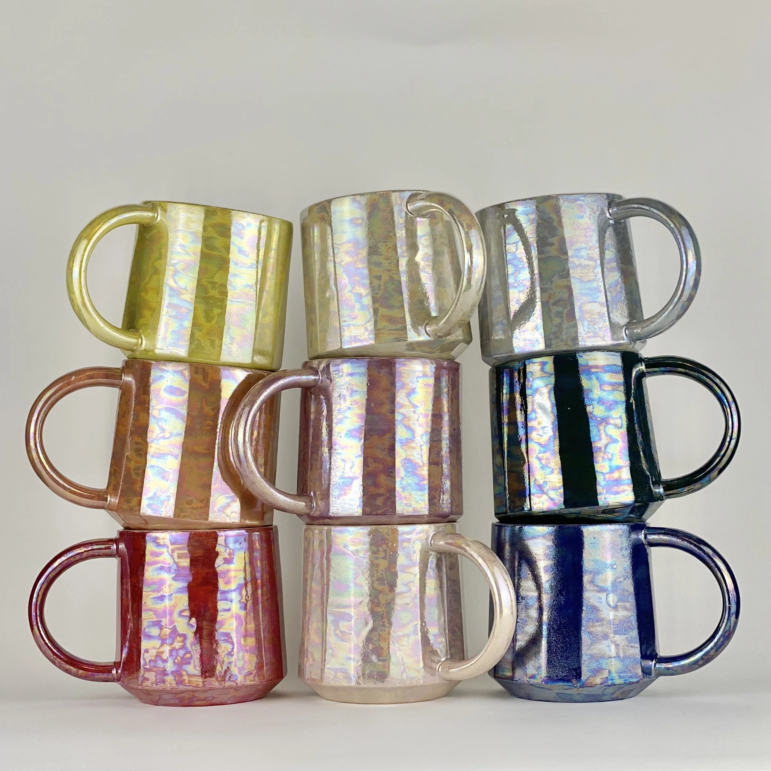 Stacked iridescent mugs in yellow, orange, blue, pink, white, and red colors on a white surface.