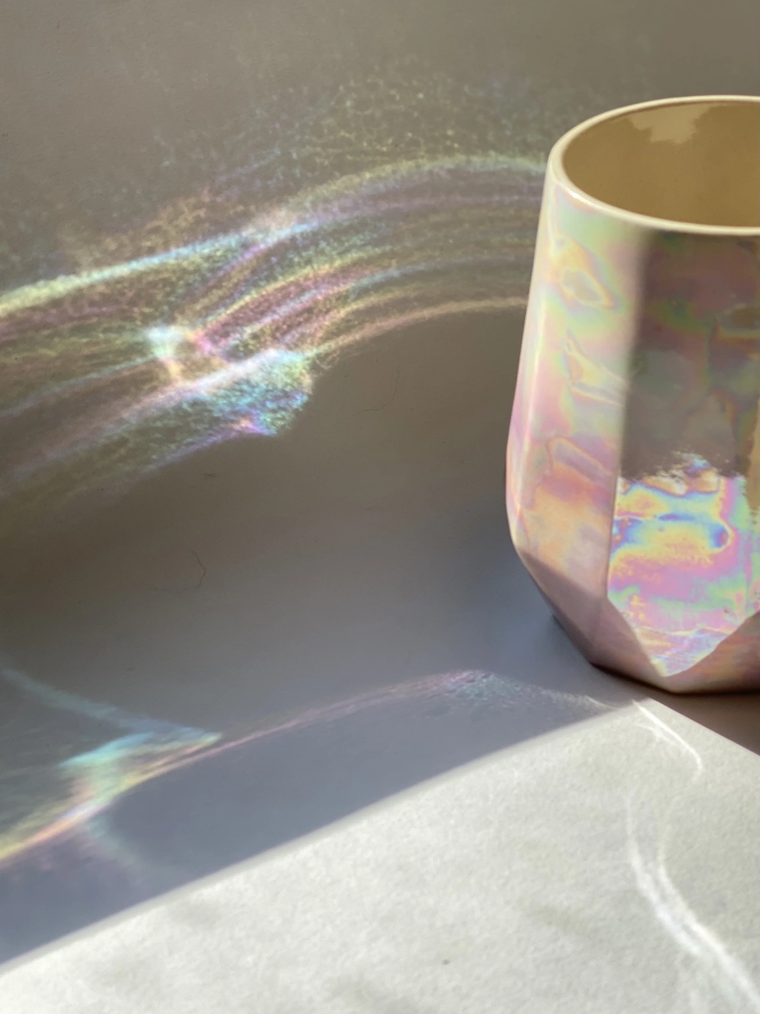An iridescent ceramic tumbler on a white surface with rainbow light reflections cast from the light hitting the iridescent glaze. 