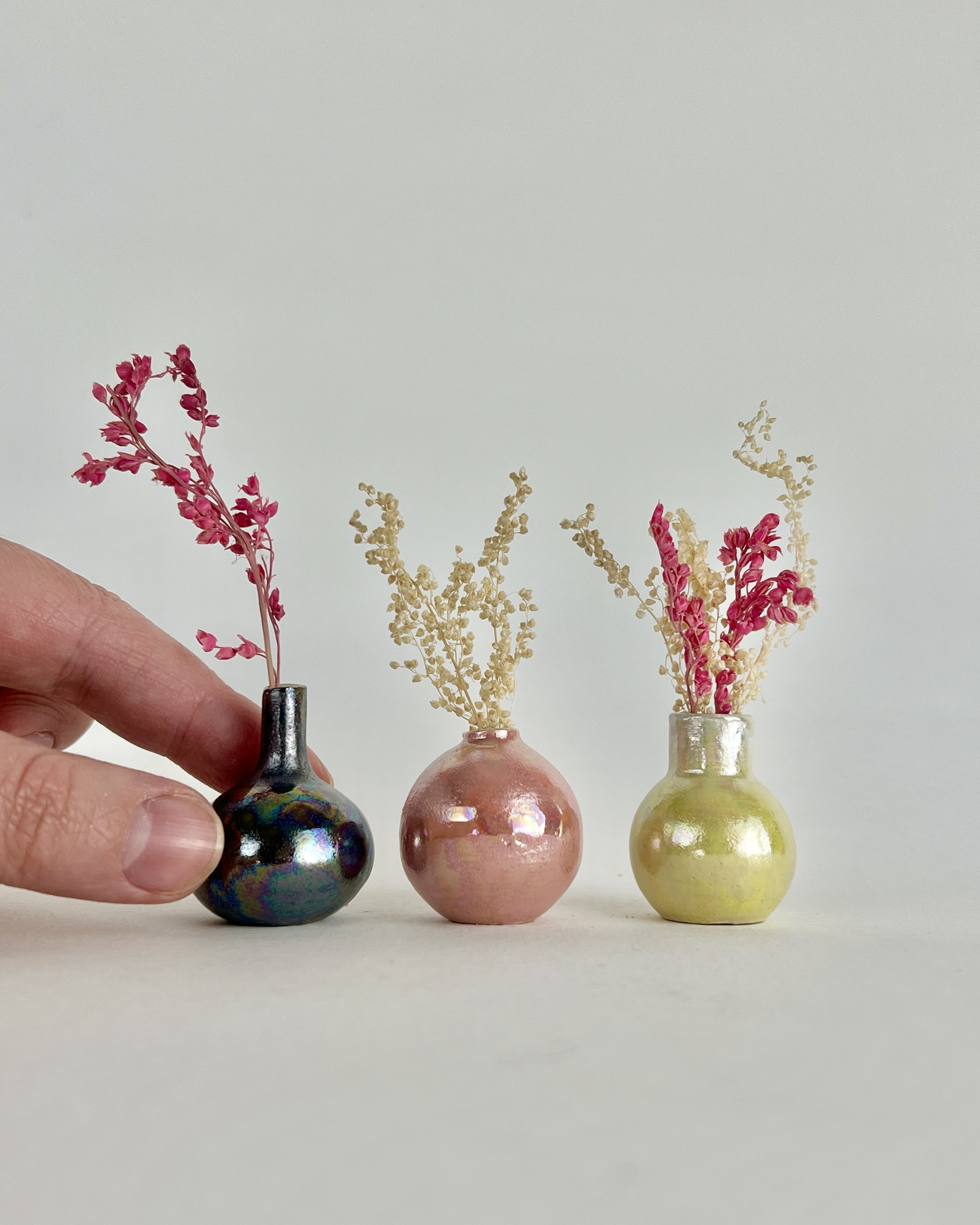 Three small decorative vases with dried flowers, one dark iridescent, one pink, and one yellow, arranged on a white surface against a plain white background, with a hand adjusting the dark vase.