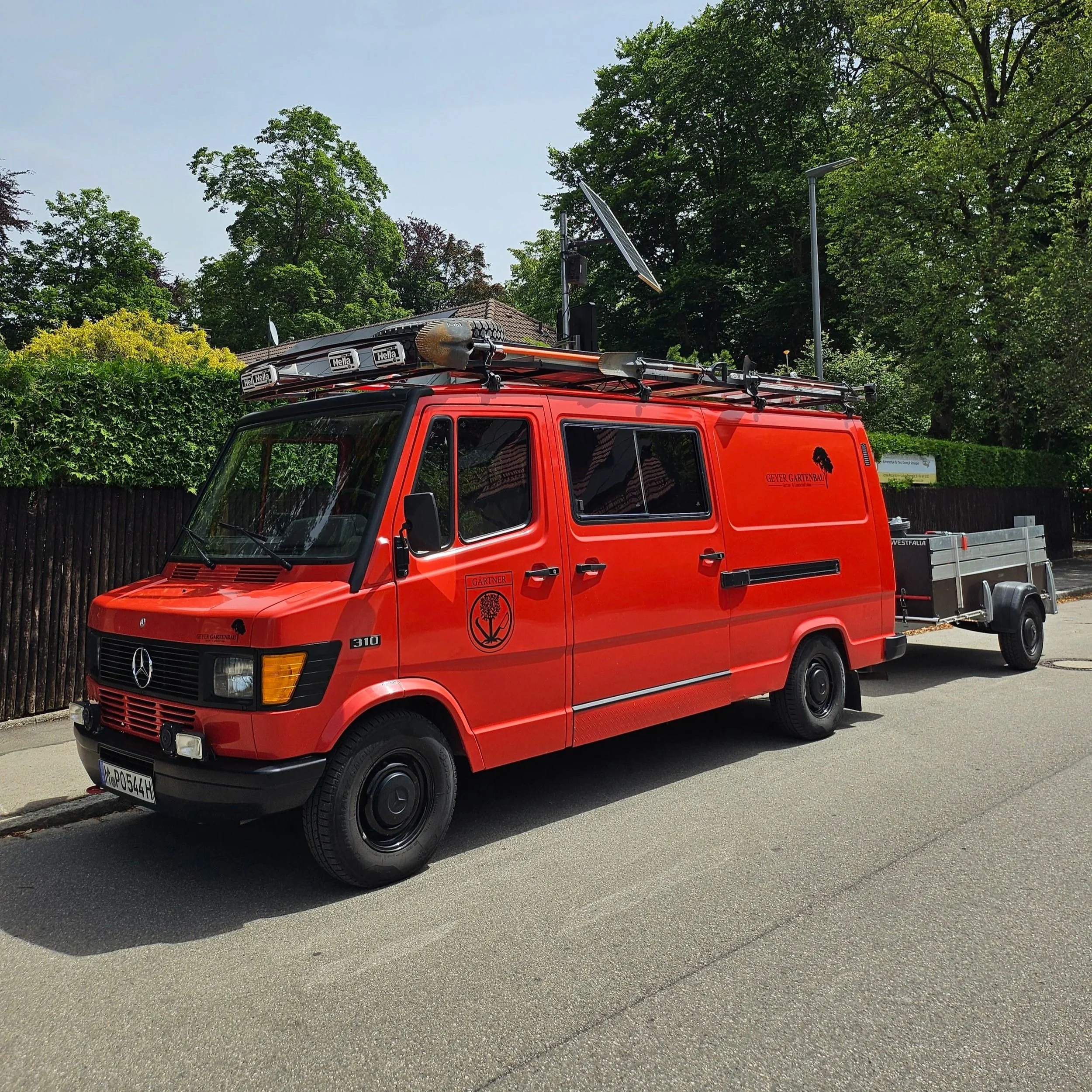 Roter Mercedes-Benz Feuerwehrfahrzeug mit Dachausrüstung und Anhänger auf einer Straße, umgeben von Bäumen und einem Zaun.