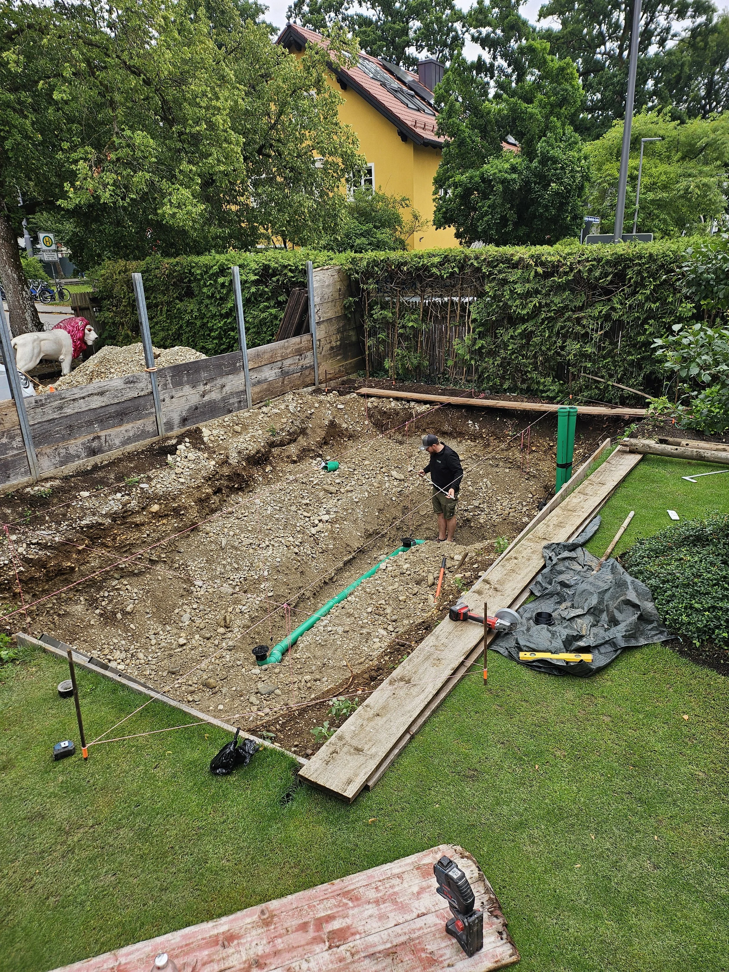Eine Baustelle im Garten, auf der eine Person gerade grabt. Es gibt Bauwerkzeuge, Holzplanken, eine große grüne Rohrleitung und eine Plane. Im Hintergrund sind Bäume, ein Zaun, ein Haus und eine weiße Ziege mit einem roten Schal sichtbar.