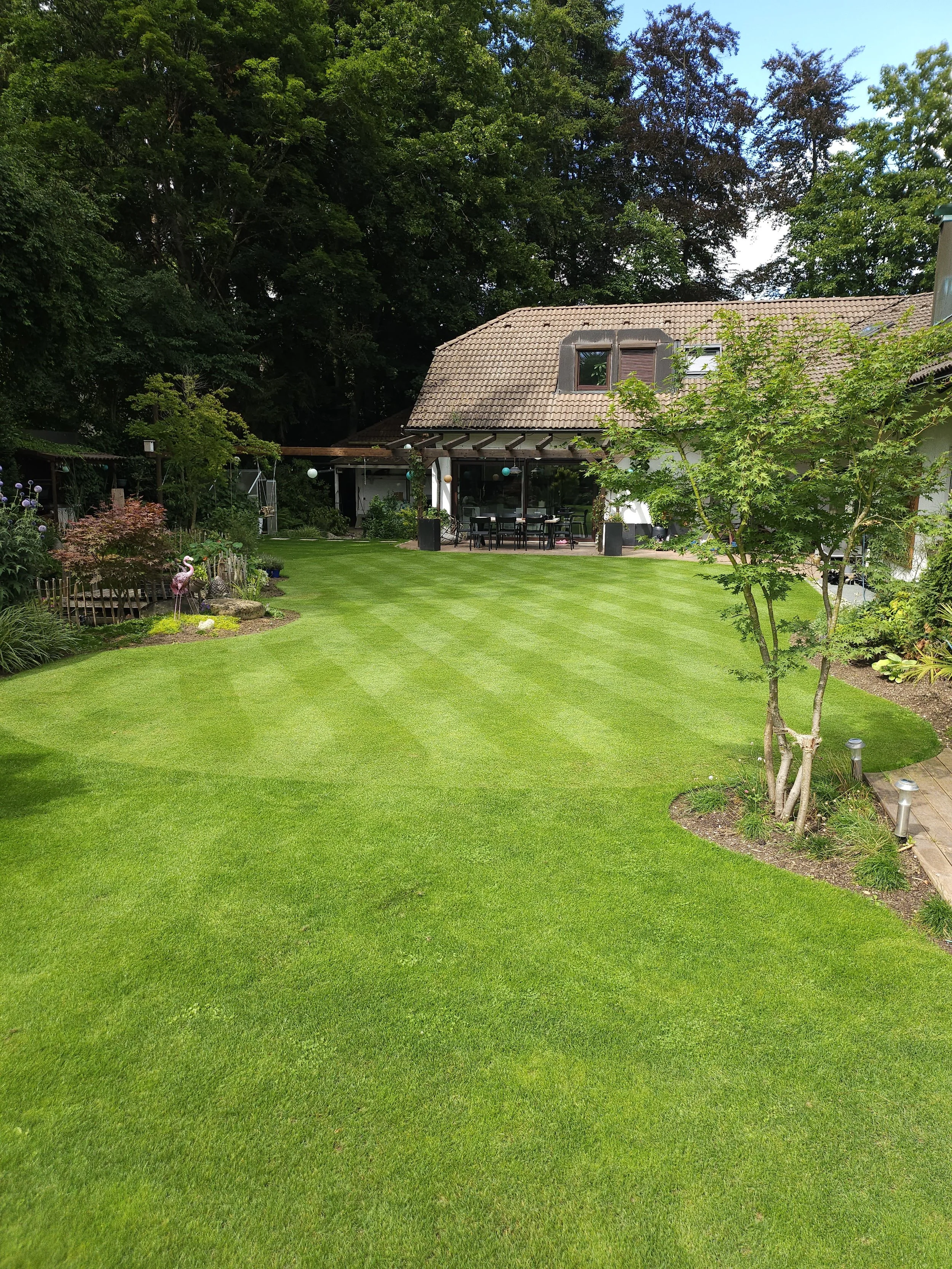 Garten mit grünem Rasen, Bäumen, Pflanzen, Gartenmöbeln und einem Haus im Hintergrund.