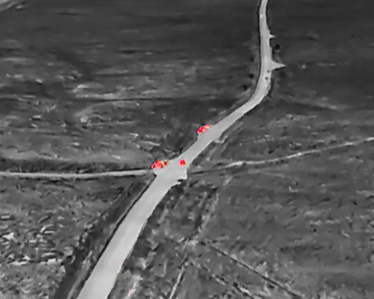 Greyscale thermal drone image of a rural road with red heat signatures indicating vehicles or persons, used for security and search and rescue (SAR) monitoring.