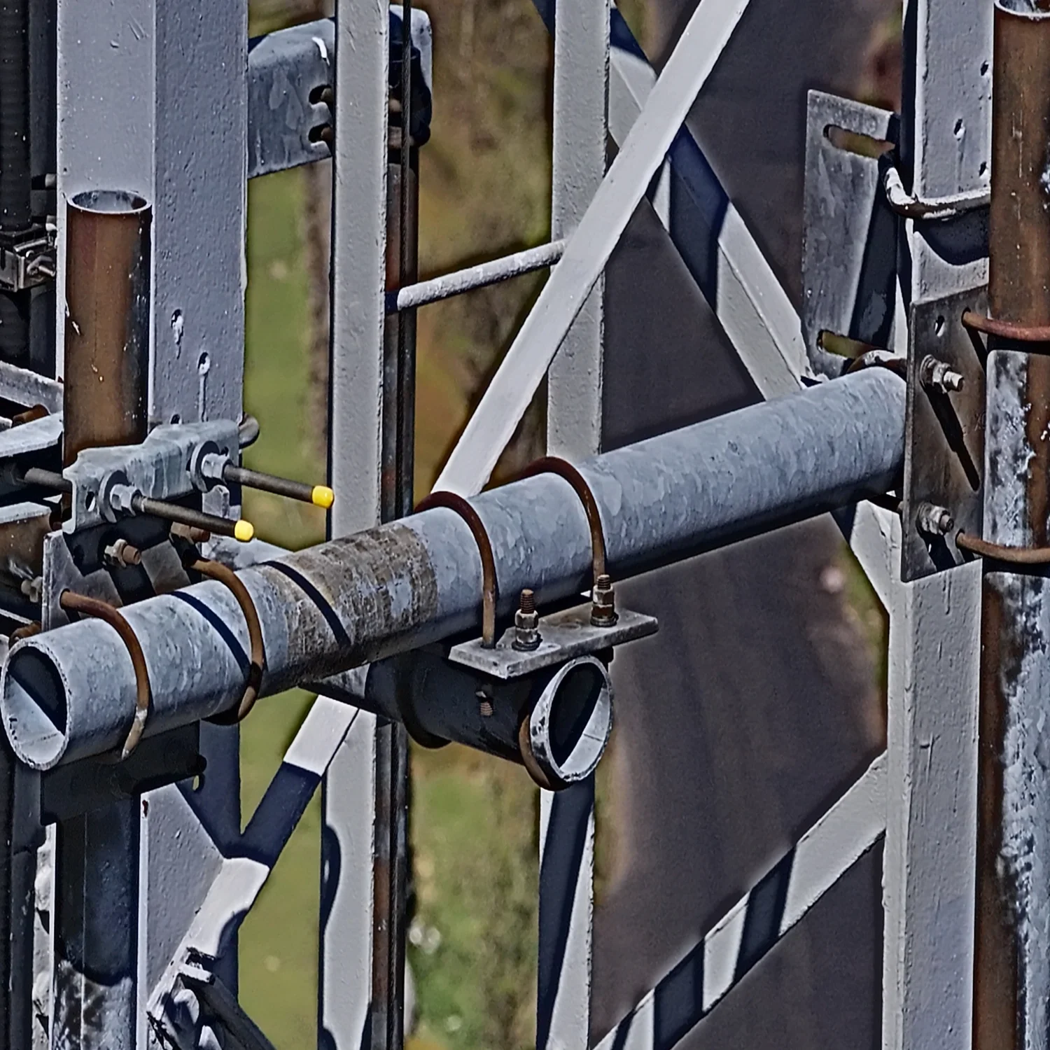 Detailed aerial drone photograph of telecommunications tower structural mounts, U-bolts, and pipework showing surface weathering and mechanical connections.