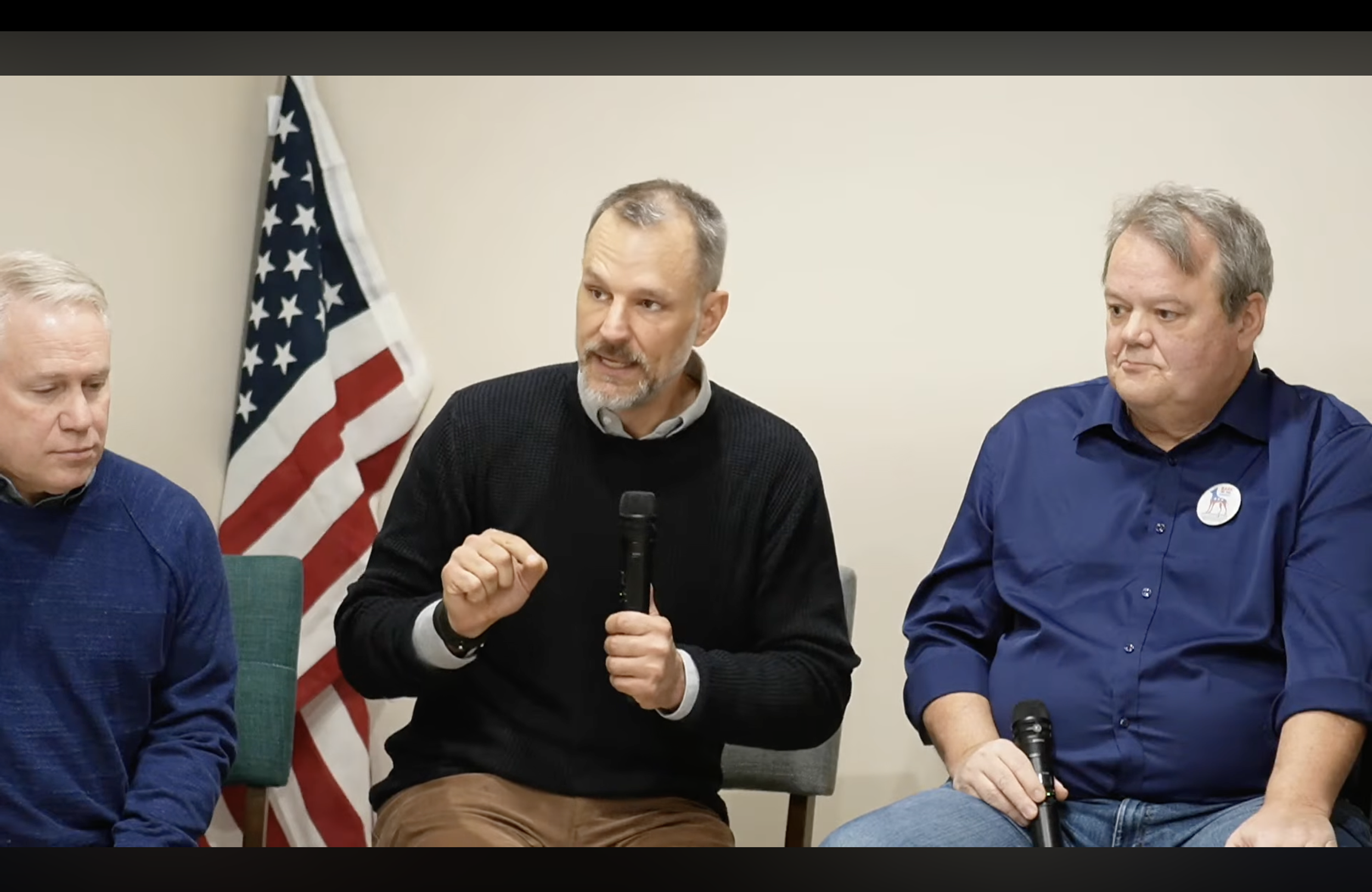 Three men sitting at a panel discussion with American flag in the background. The man in the middle is speaking into a microphone, wearing a black sweater. The man on the right has a badge on his blue shirt and is holding a microphone.