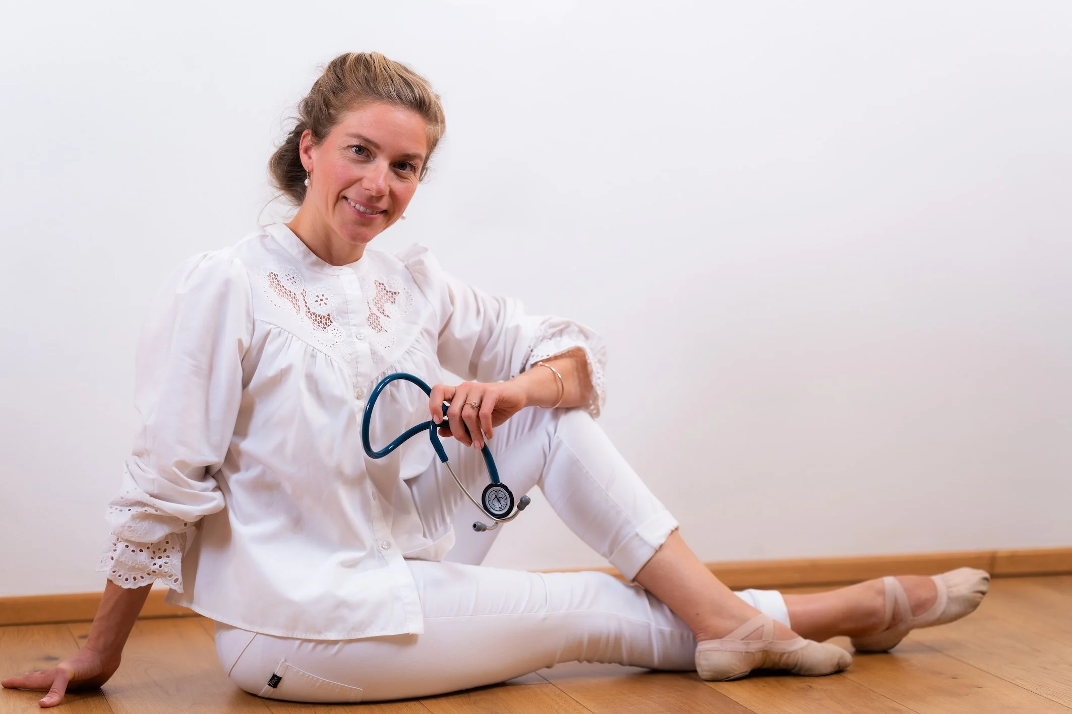 Eine Frau in weißem Kleid mit einem Stethoskop, die in einer Yoga-Position auf einem Holzfußboden sitzt, gegen eine weiße Wand.