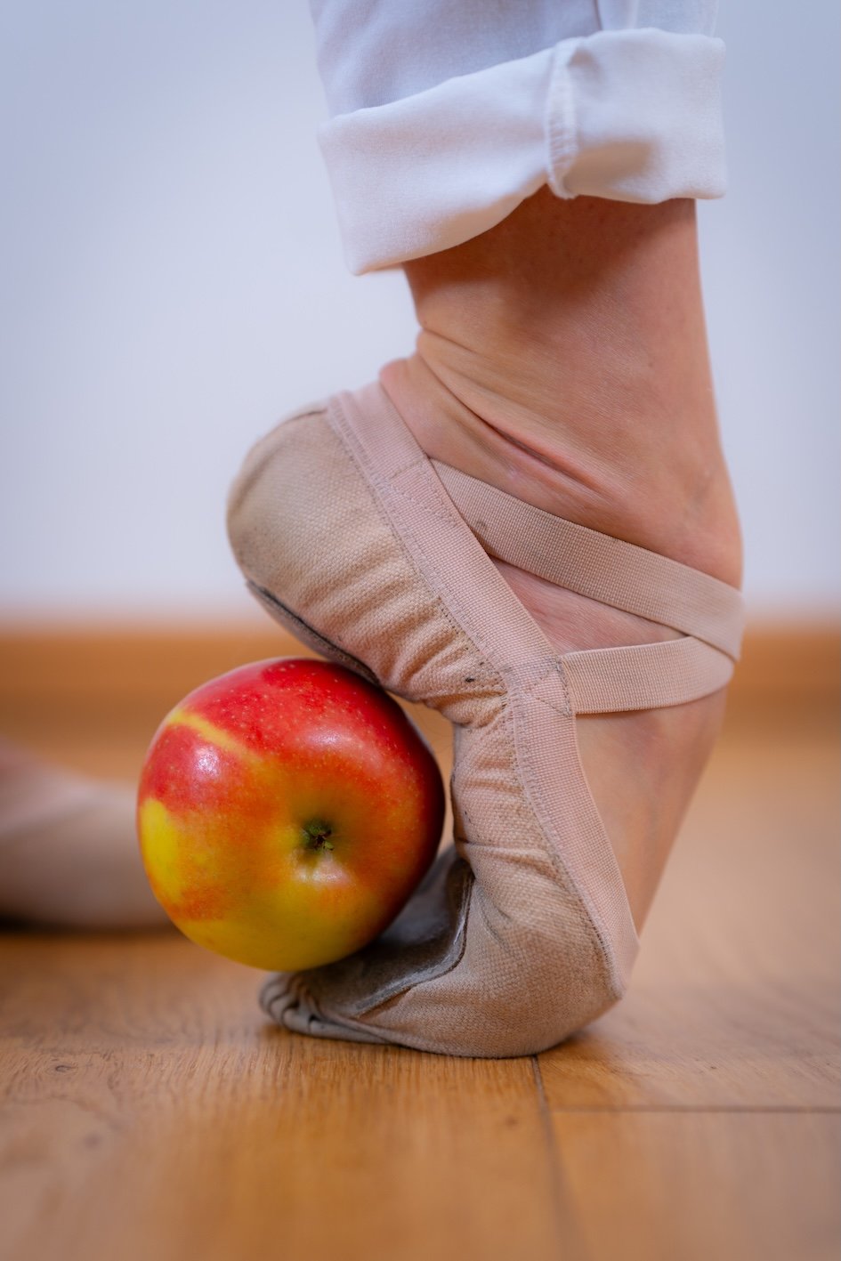 Eine Person sitzt auf einem Boden, deren Fuß ist mit einem Zehensteg-Schuh bedeckt, der an einem Apfel befestigt ist, der auf dem Boden liegt.