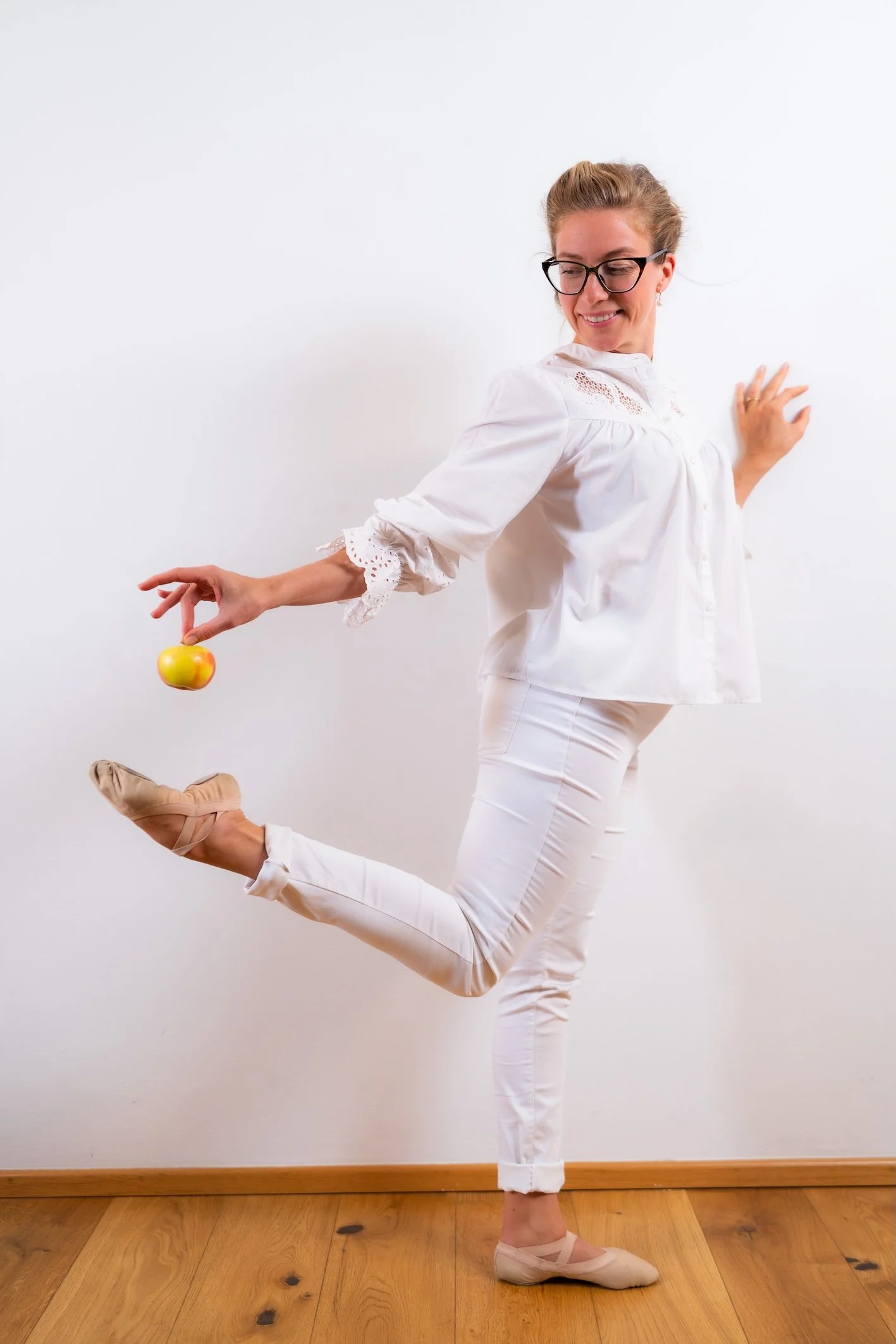 Frau in weißen Kleidung führt Ballettübung vor, steht auf einem Bein, das andere hinter ihr gekreuzt, auf einem Holzfußboden vor weißer Wand.
