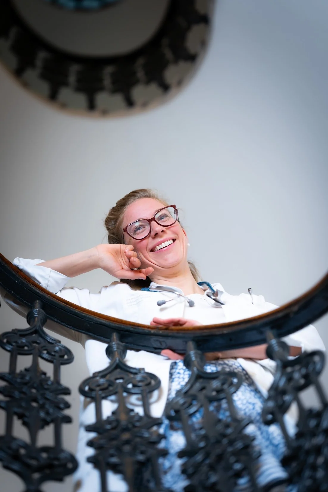 Blick in einen Spiegel zeigt eine lächelnde Frau in einem weißen Arztkittel mit Brille und Stethoskop. Der Spiegel hat ein dekoratives schwarzes Gestell.