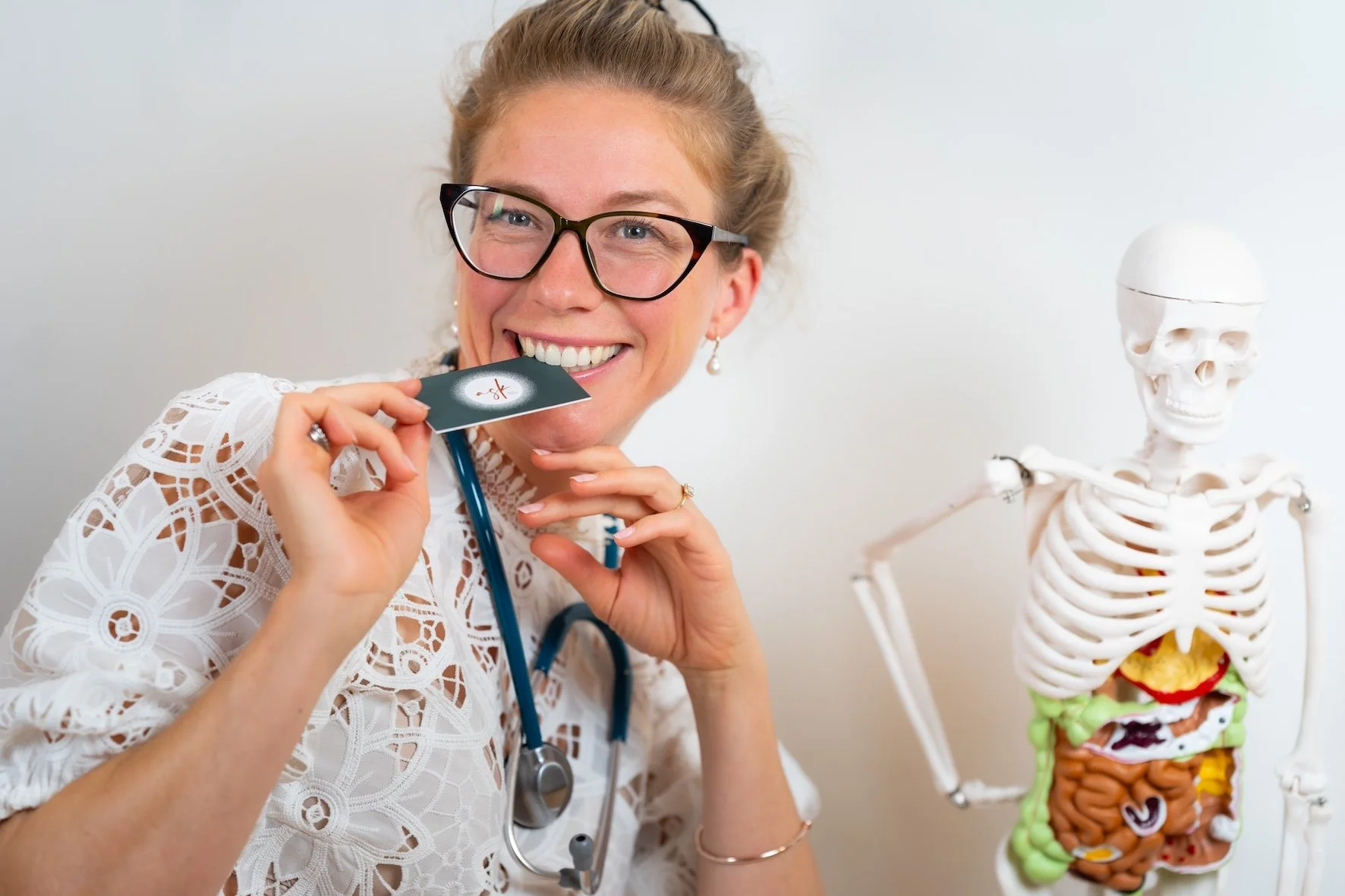 Eine lächelnde Frau in einem weißen Spitzenkleid trägt eine Brille und hält ein Stethoskop. Neben ihr steht ein plastischer menschlicher Anatomie-Model mit einem offenen Brustkorb, der die inneren Organe zeigt.