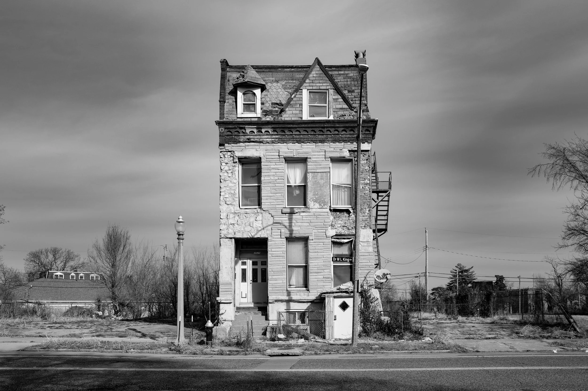 Black and white photo of an old, dilapidated three-story house with boarded-up windows, a fire escape on the side, and a crooked mailbox in front, on a deserted street with bare trees and a cloudy sky.