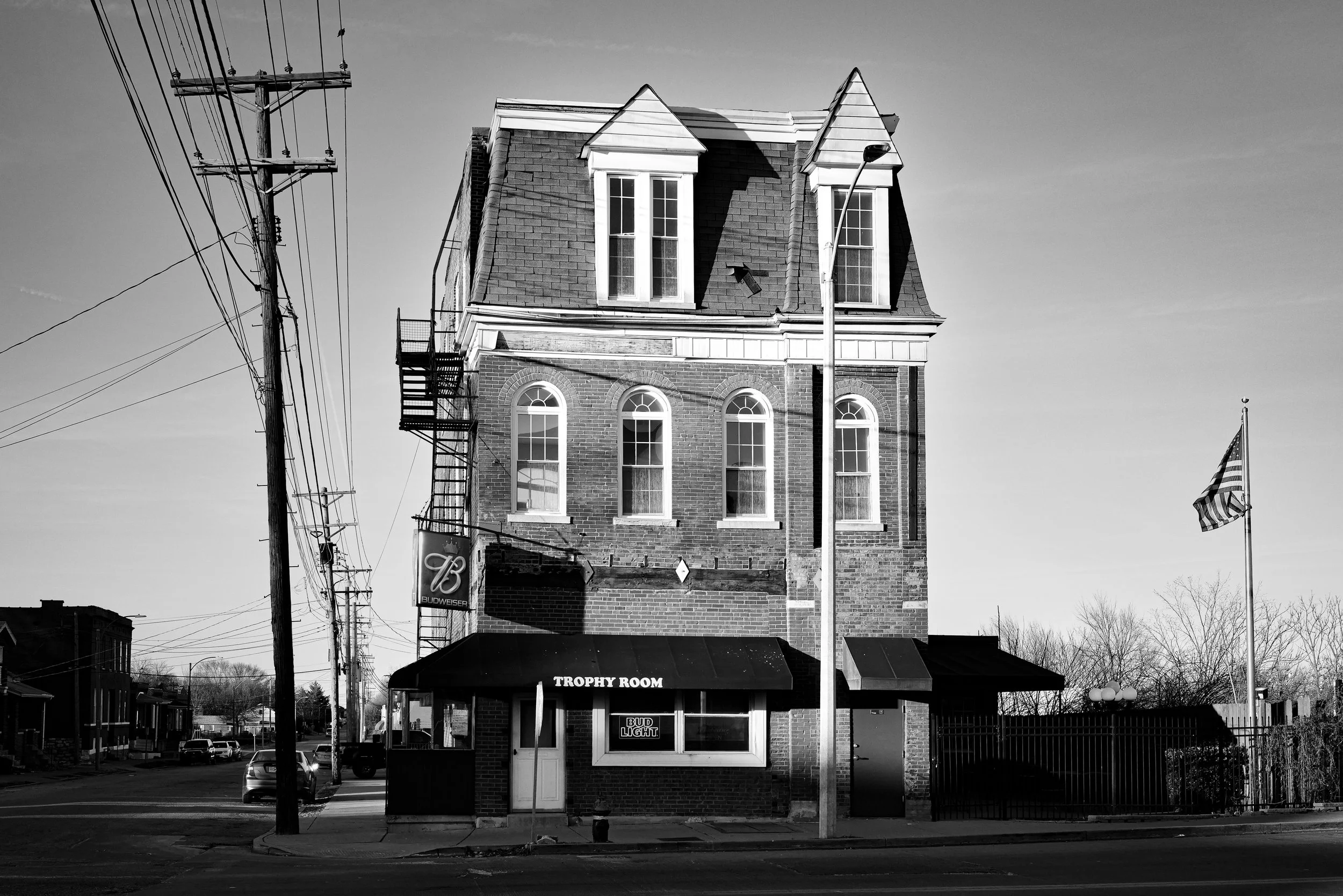 A black and white photo of a three-story brick building with multiple windows, a near corner store with an awning labeled 'TROPHY ROOM,' a sign for Budweiser, and an American flag on a pole to the right, adjacent to electric poles and wires.