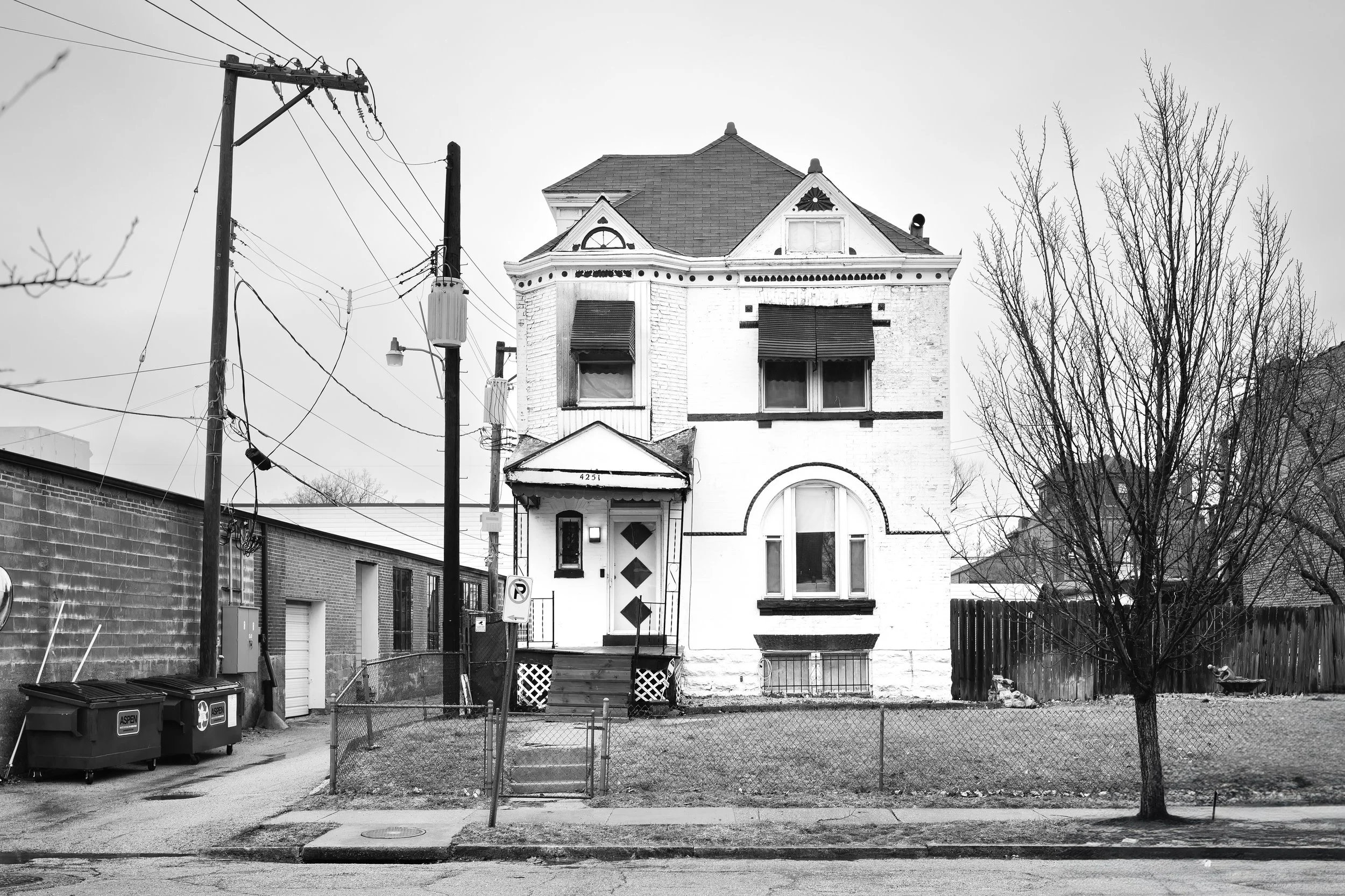 A black-and-white photo of a worn, two-story house with a small front porch, situated behind a chain-link fence, with leafless trees and utility poles nearby.