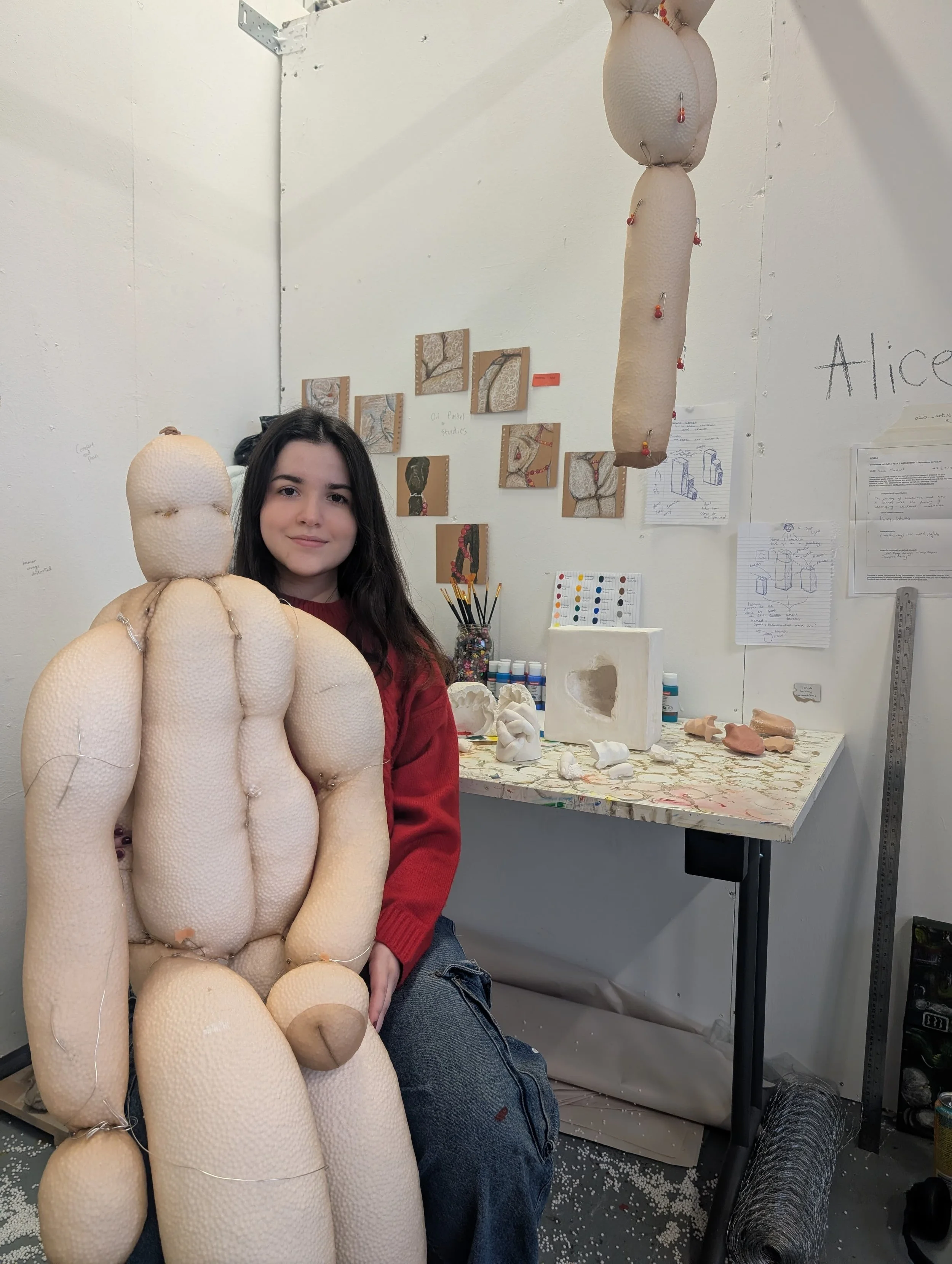 Young woman sitting in an art studio holding a large, handmade fabric sculpture resembling a seated humanoid figure. The studio has a white wall with artwork, art supplies on a table, and hanging foam sculptures.
