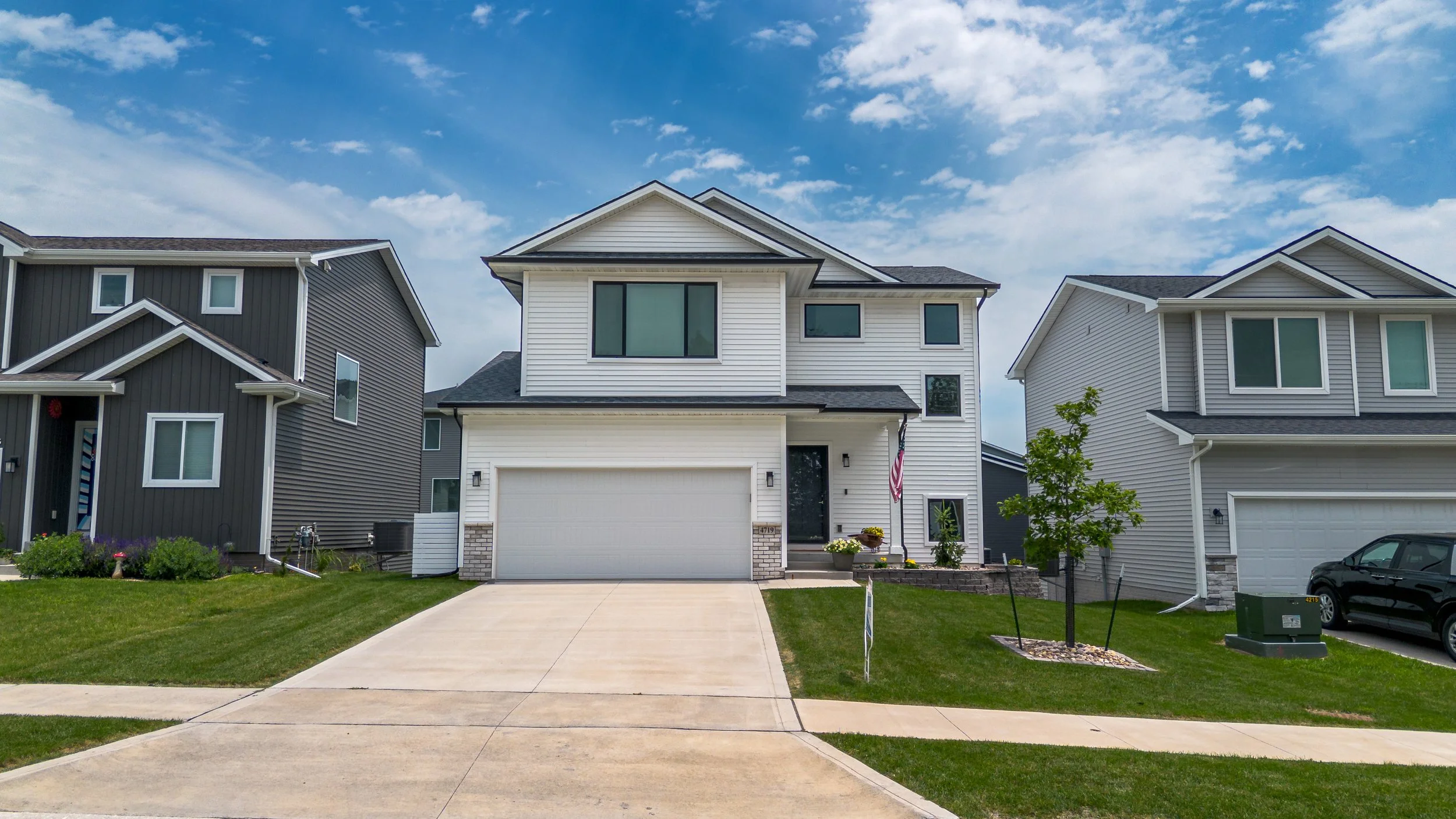 A modern white two-story house with a garage, small front yard, and well-maintained lawn, situated between two other similar houses under a partly cloudy sky.