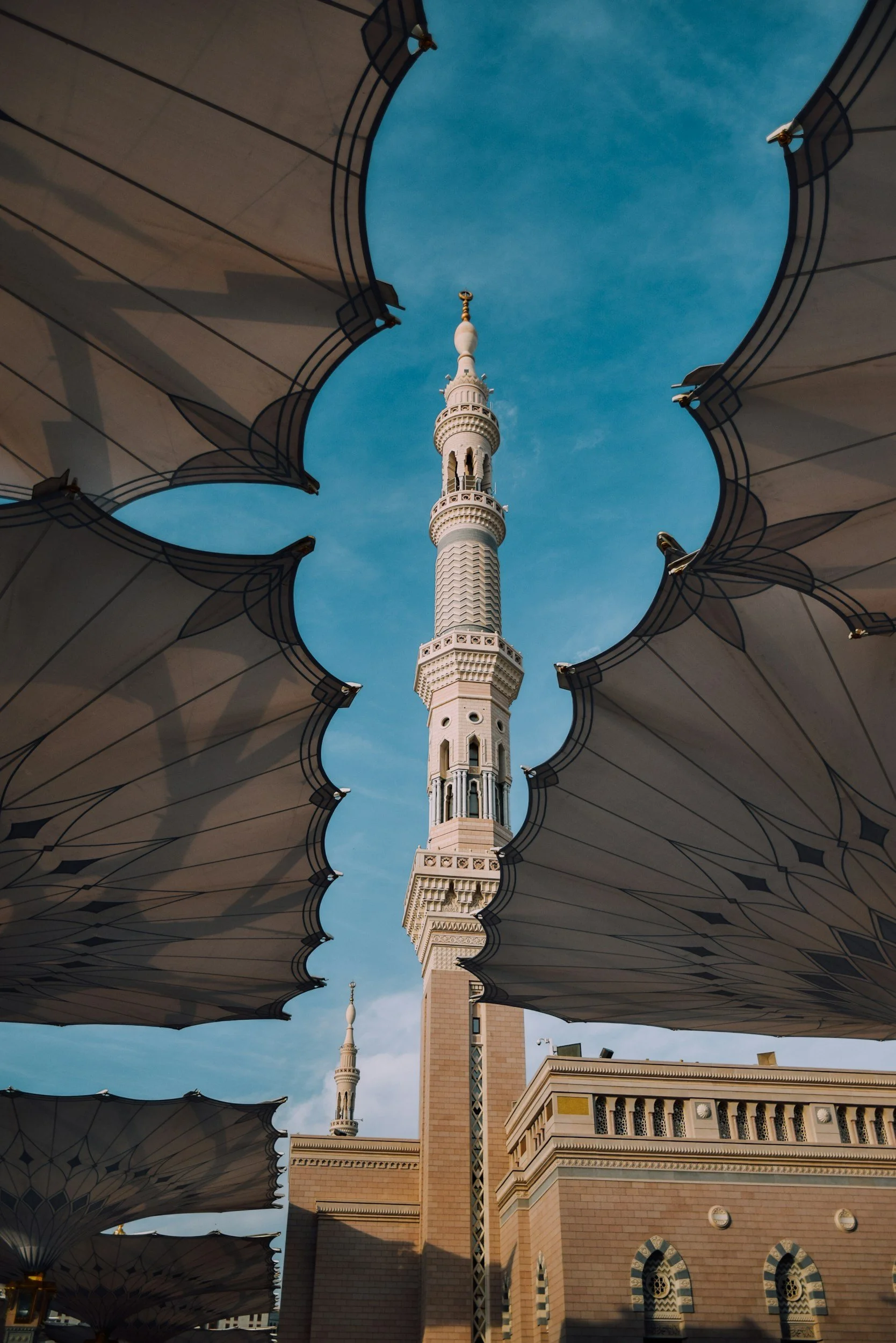 A tall, ornate Muslim minaret viewed through decorative umbrellas, with a blue sky background.