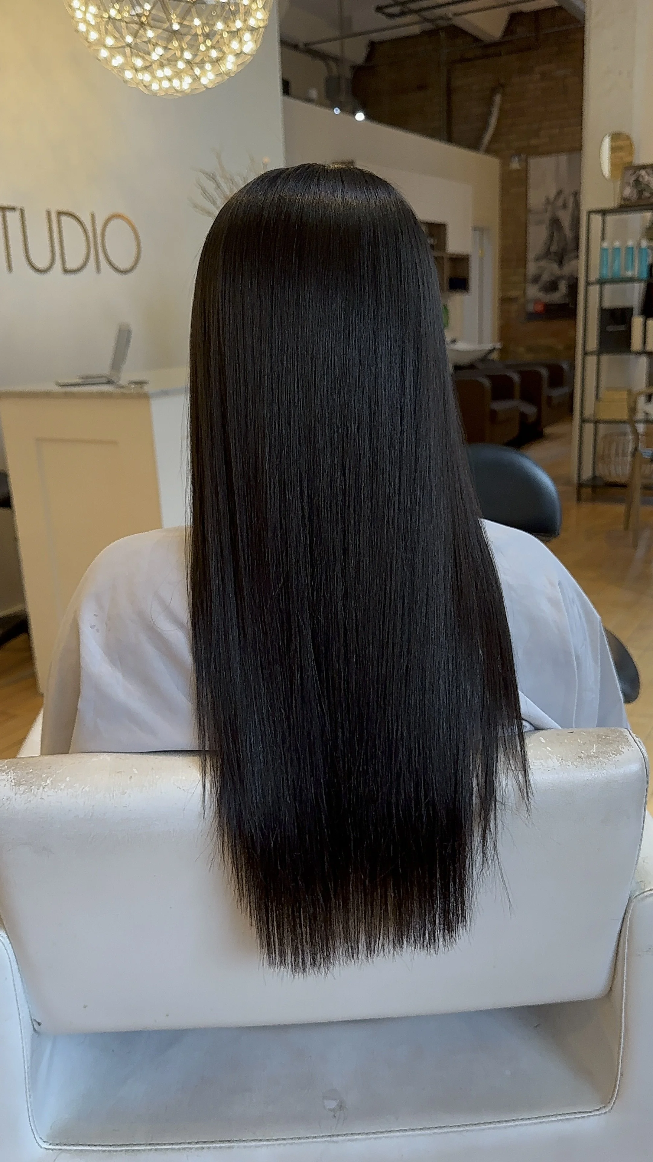 Woman with long, straight black hair sitting in a salon chair, showing the hair from the back. The hair is shiny and well-maintained. The salon has a modern and cozy interior with a chandelier, shelves, and a sign that says 'STUDIO'.