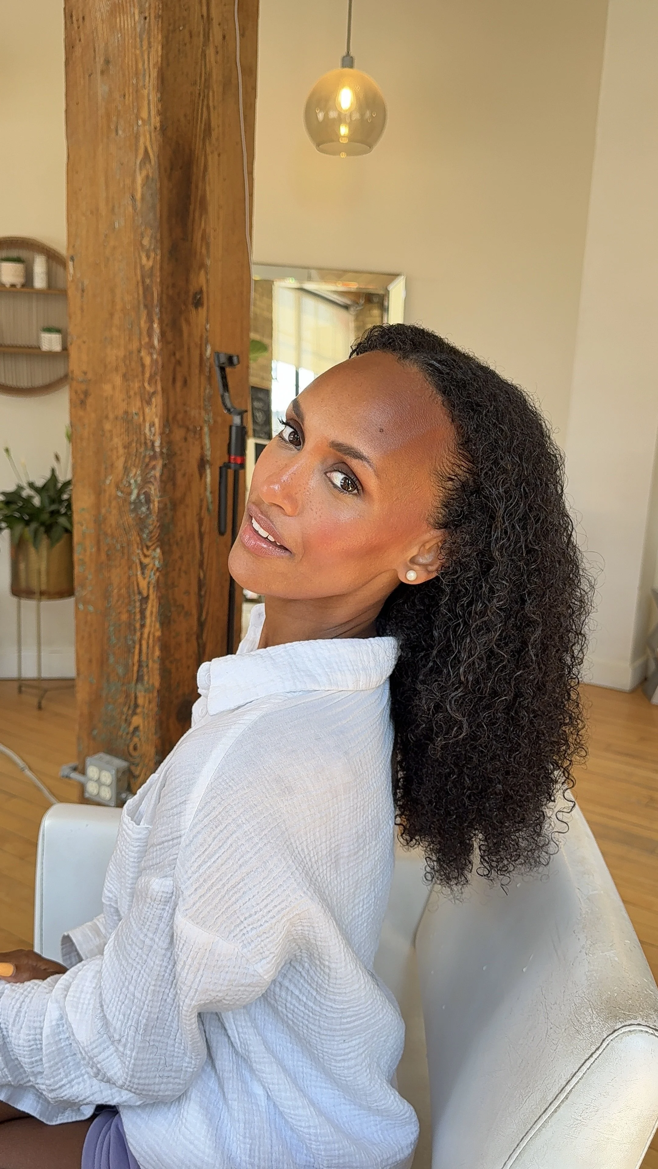 A woman with curly hair wearing a white shirt, sitting on a white chair in a room with wooden floors and a wooden beam. She is looking over her shoulder at the camera with a slight smile, in a well-lit room with modern decor.