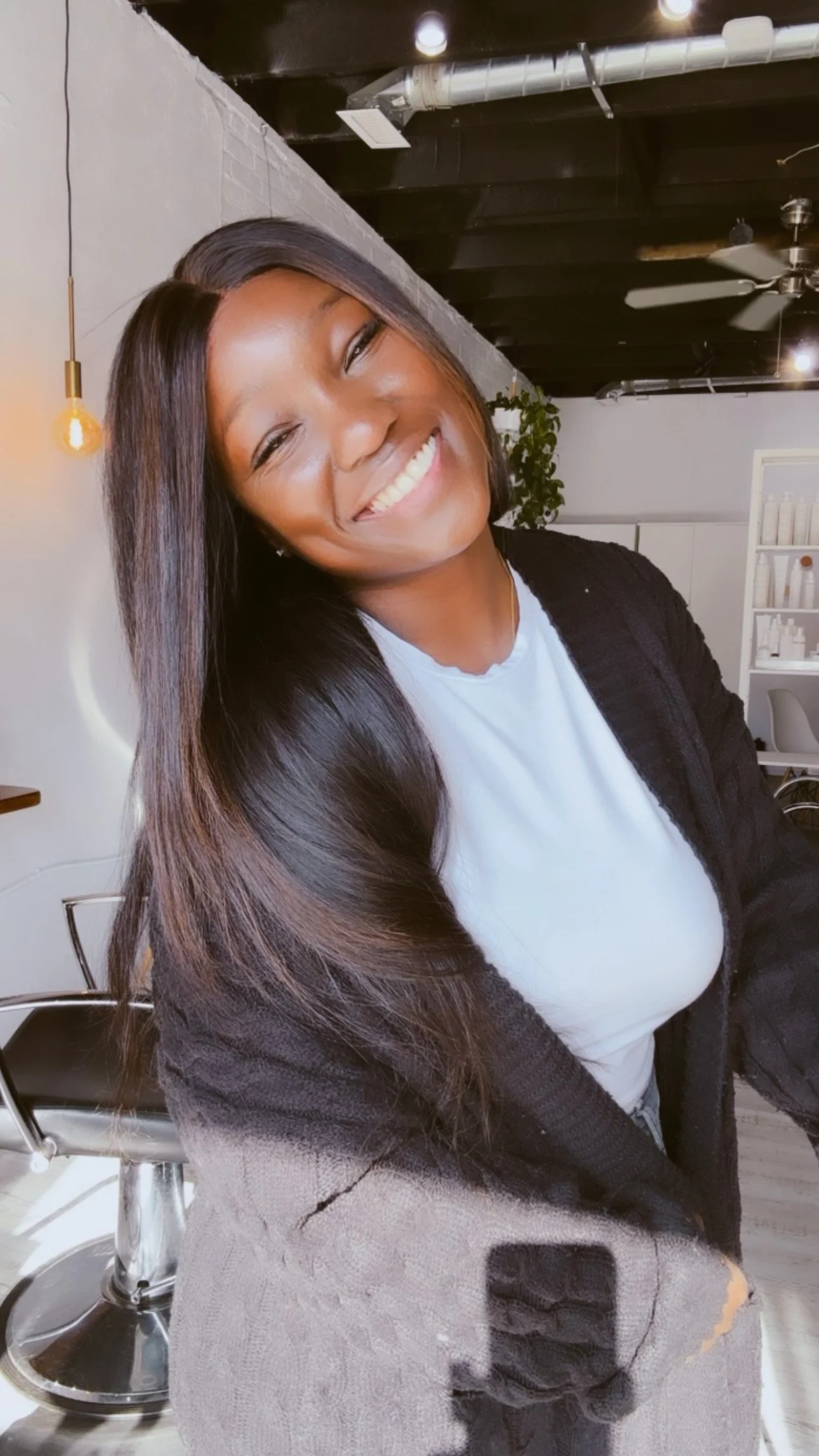 A woman smiling with long dark hair, wearing a white shirt and a black jacket, sitting in a modern space with a white wall, shelf, and ceiling fan.