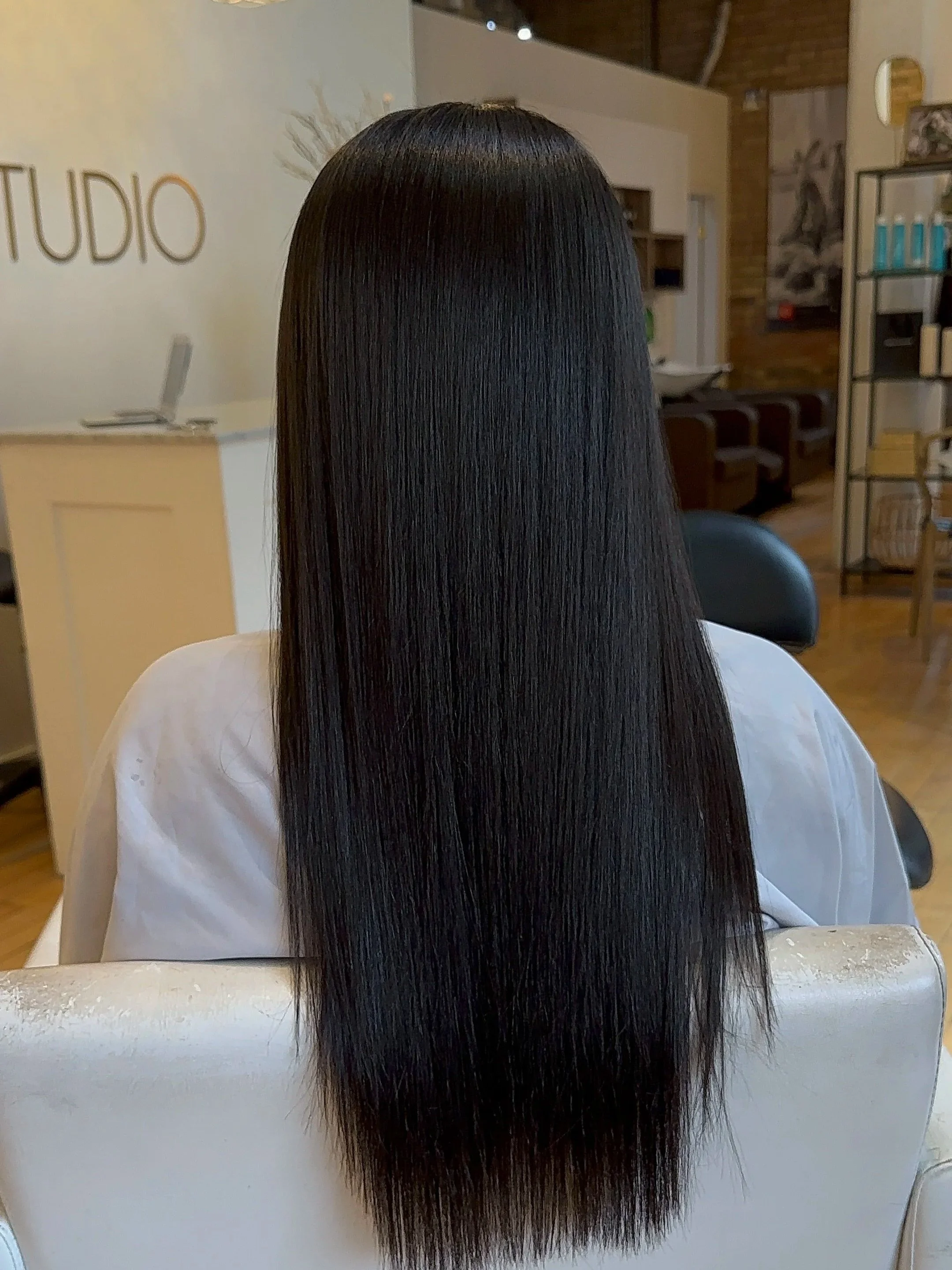 Back of a person with long, straight, dark hair in a salon, sitting in a chair.