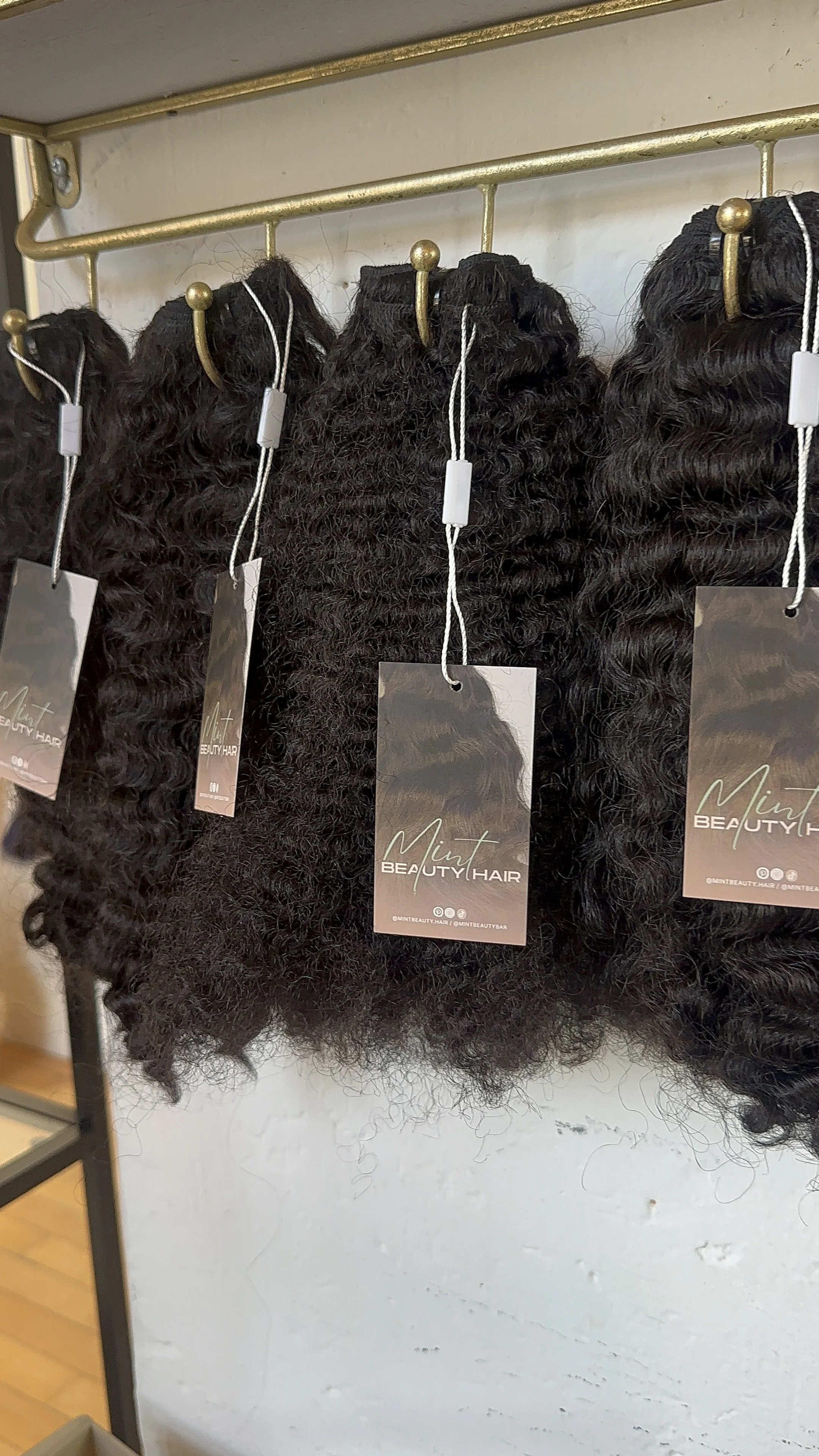 Bundles of curly black hair extensions hanging on a wall with tags that say 'Mint Beauty Hair'.