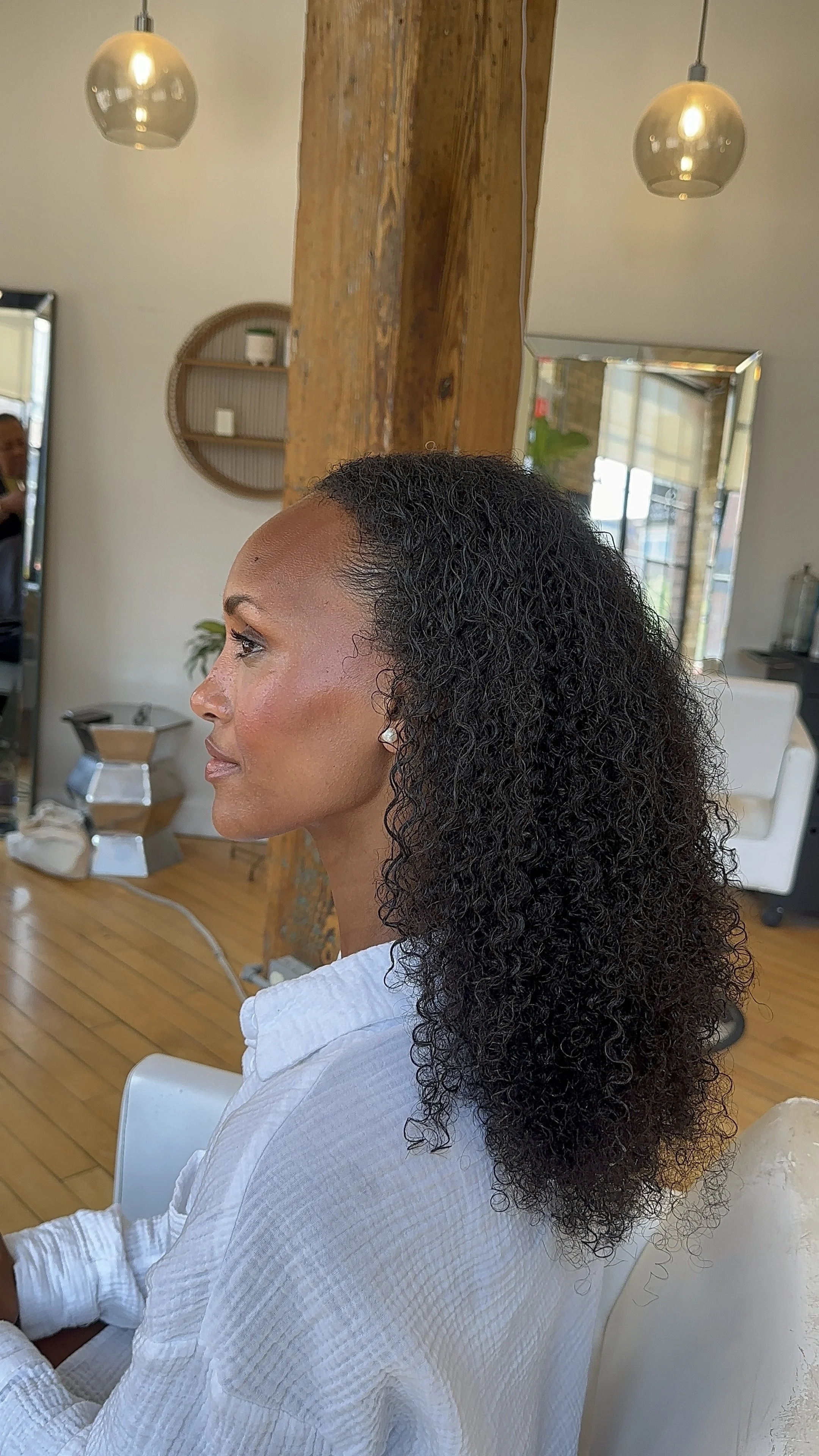 A woman with curly black hair sitting in a salon, facing left, with a peace sign necklace, wearing a white textured shirt. The salon has wooden floors, a wooden beam, hanging pendant lights, and a mirror reflecting a person.