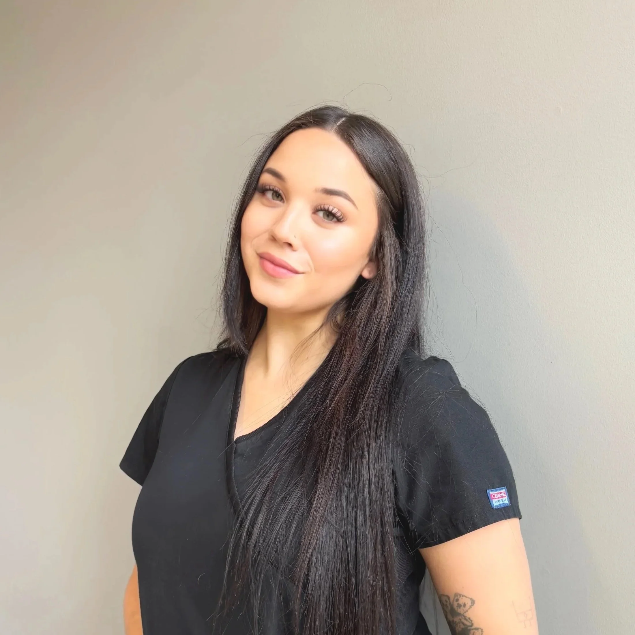 A young woman with long, dark hair and light makeup, wearing a black medical uniform, standing against a plain wall.