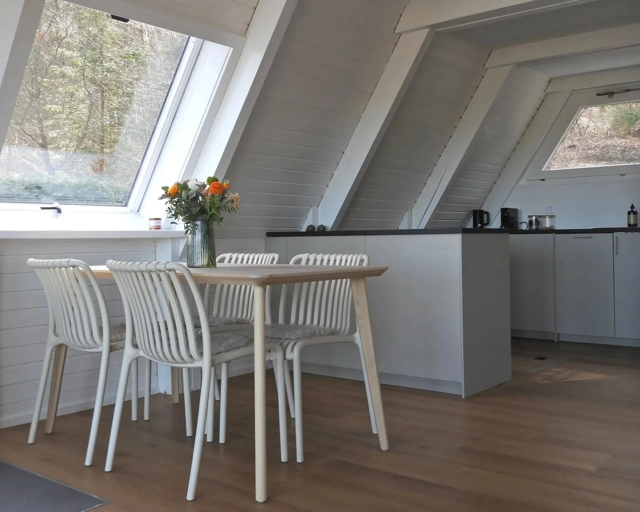 Innenaufnahme einer modernen Küche mit weißen Möbeln und großen Dachfenstern, darunter ein Esstisch mit weißen Stühlen und einem Blumenstrauß in einer Glasvase.