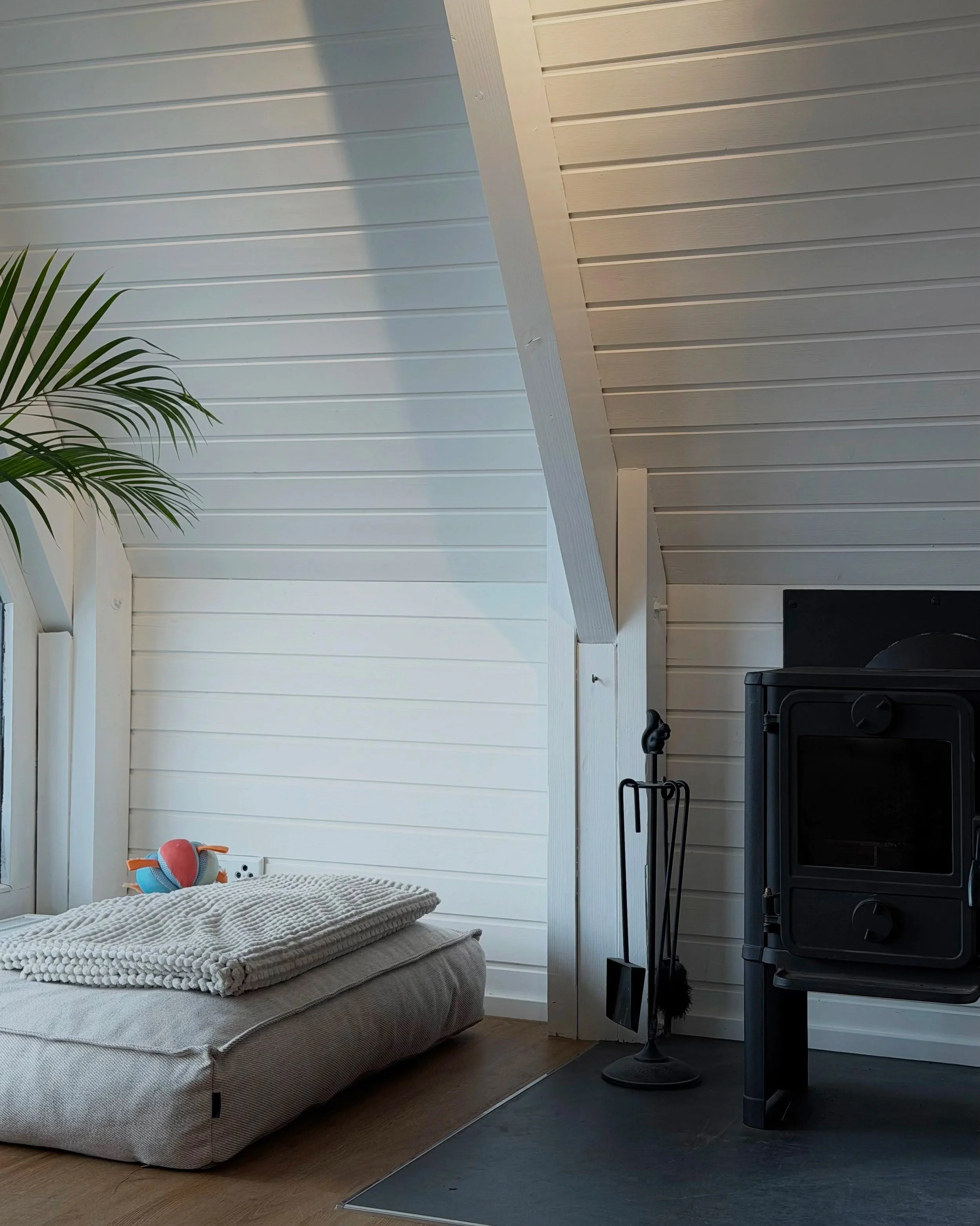 Ein Schlafzimmer mit einem Bett, einer Decke, einer Stehlampe, einer Heizung, einer Wand mit weißen Holzpaneelen und einem Fenster, das Tageslicht hereinlässt.