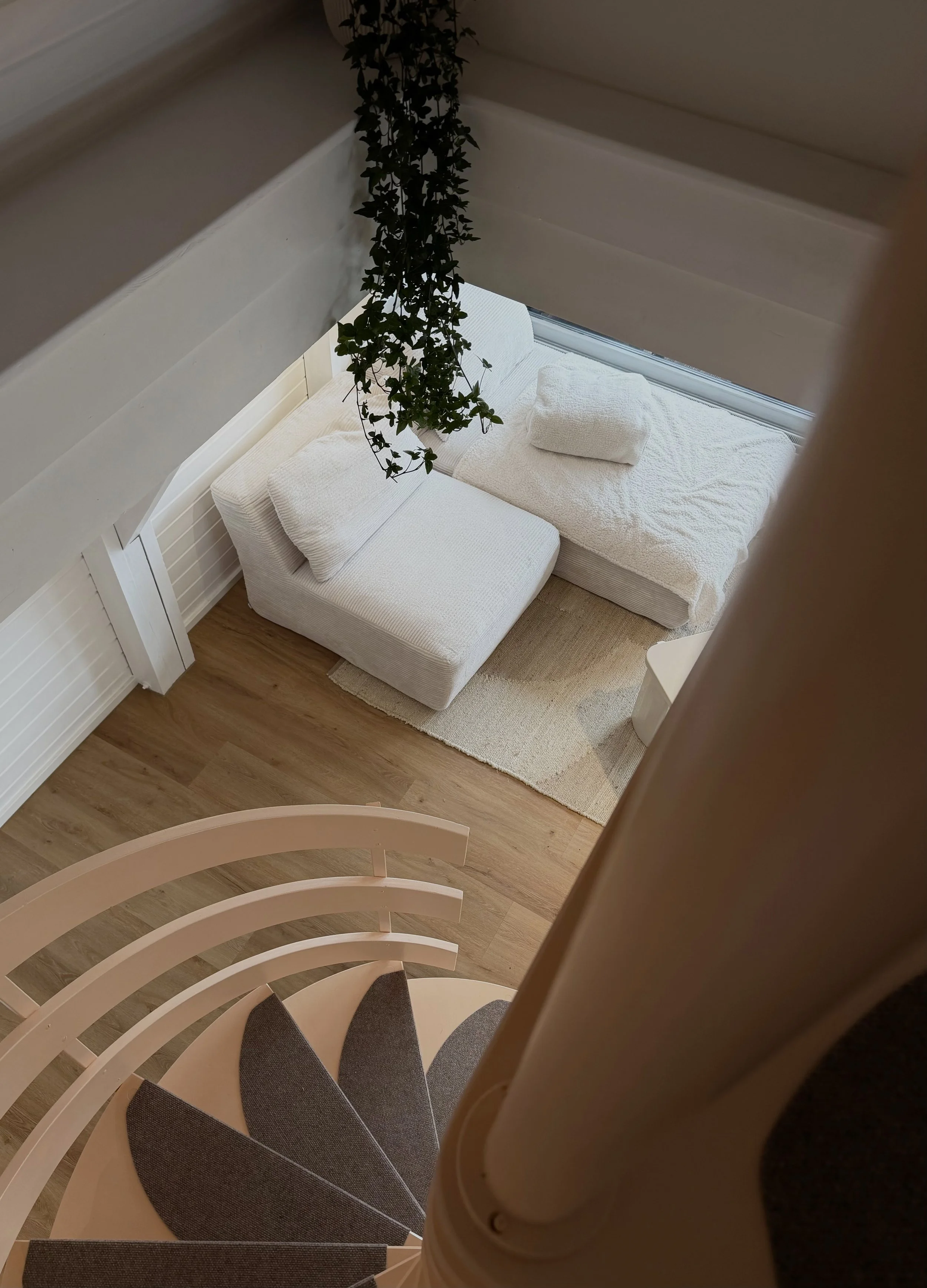 Aufstieg auf eine weiße Wendeltreppe, Blick von oben nach unten, mit grauem Teppich auf den Stufen und einer weißen Wand mit Fenster im Hintergrund, ein Sofa mit weißen Kissen in einem Raum darunter sichtbar