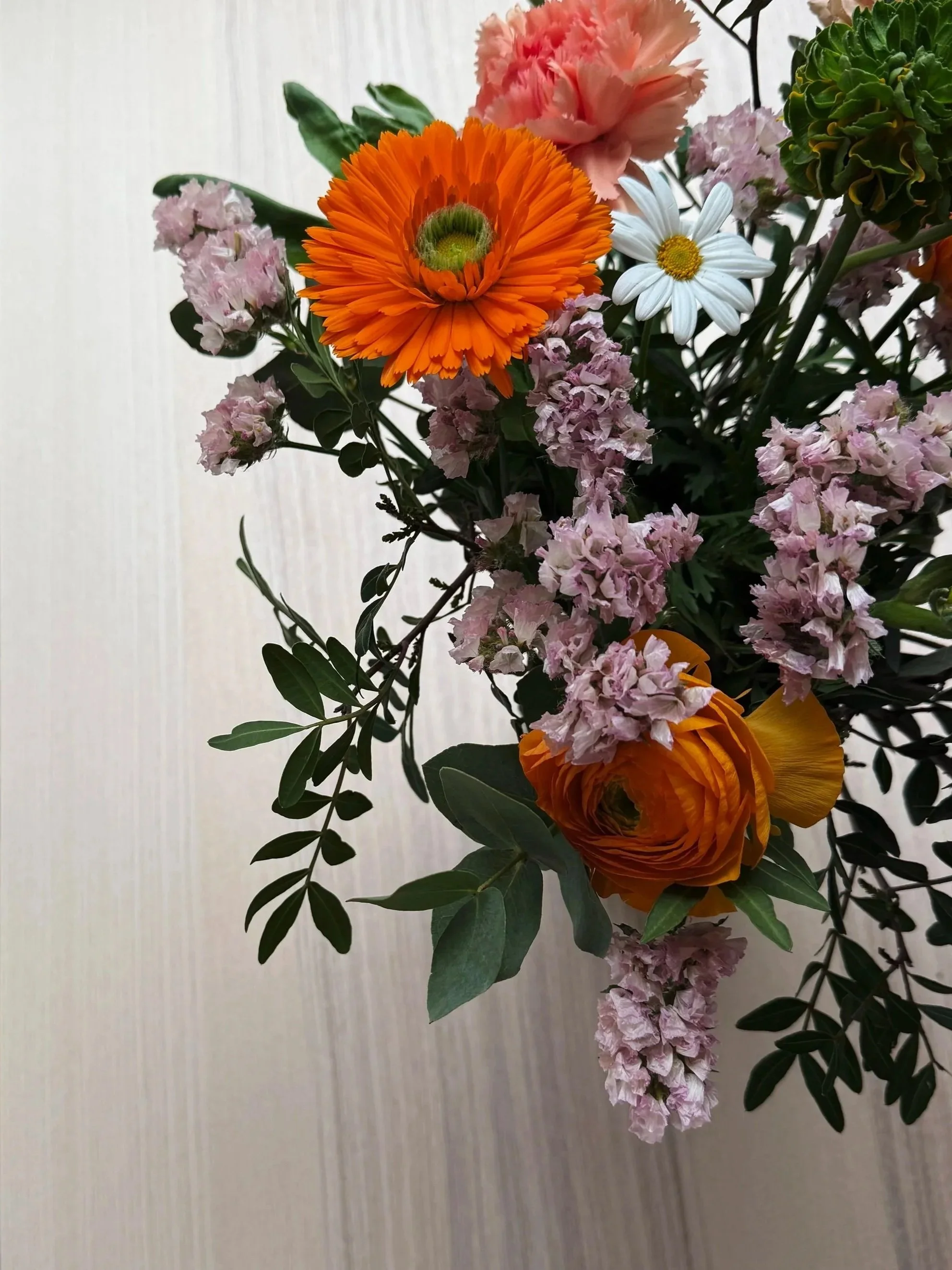 Blumenstrauß mit orangefarbener Gerbera, weißen Gänseblümchen, pinken Nelken und grünem Laub auf cremefarbenem Hintergrund.