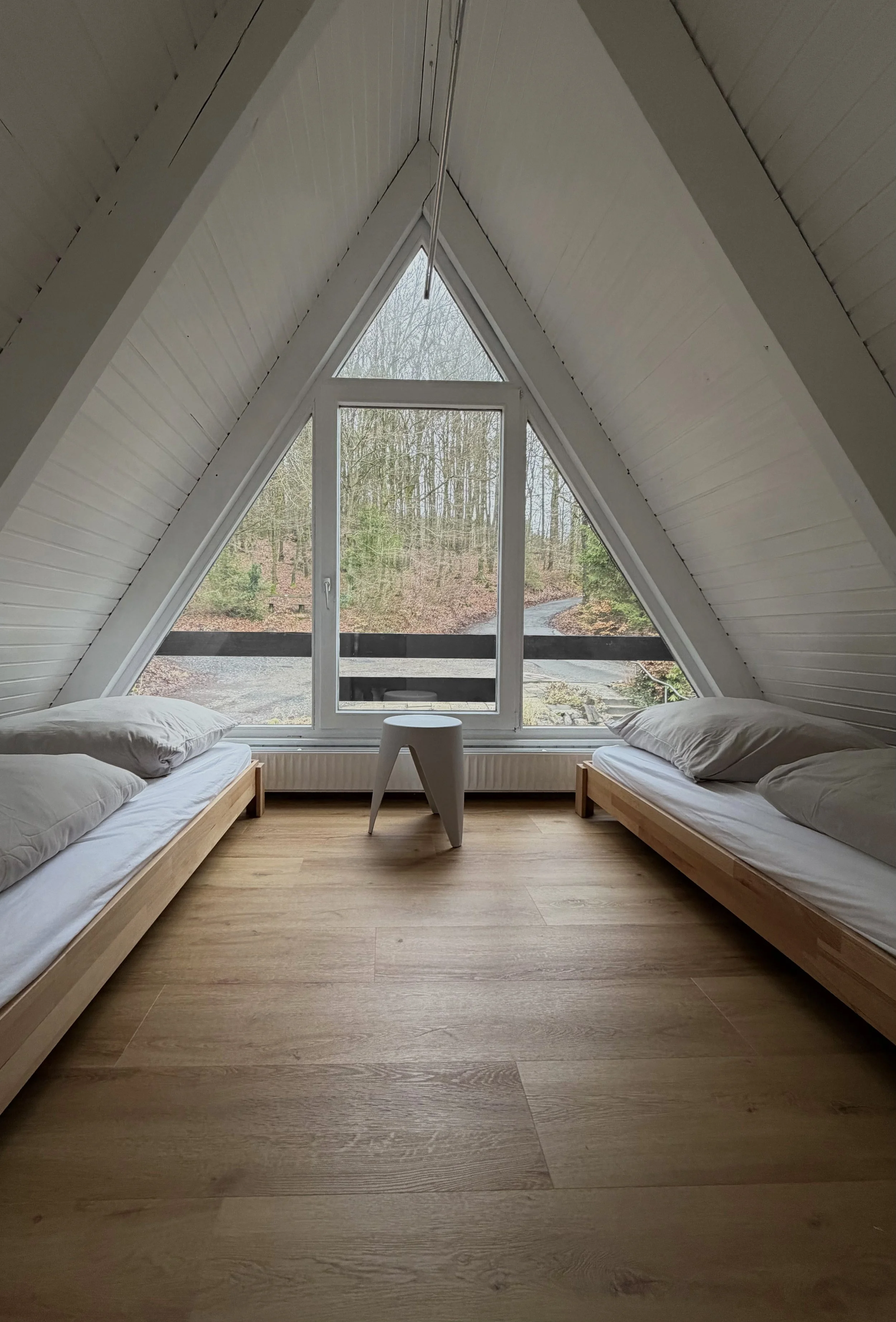 Ein Schlafzimmer mit zwei niedrigen Holzbettgestellen auf beiden Seiten, weißen Betten und Kissen, einer kleinen weißen Beistmtisch in der Mitte, großen dreieckigen Fenster mit Blick auf eine bewaldete Landschaft, und einem Dach mit weißen Paneelen.