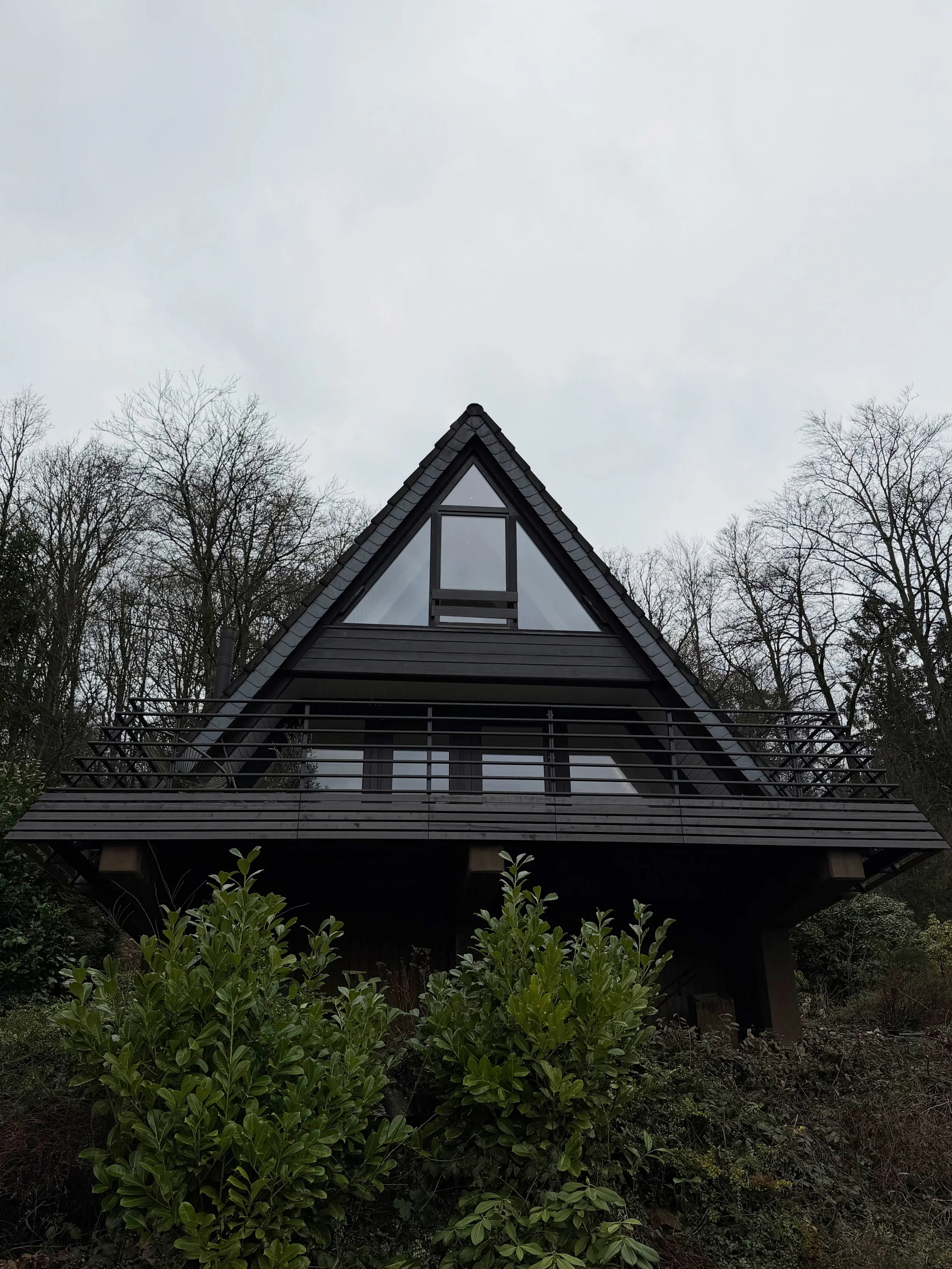 Ein modernes Haus mit einer auffälligen, schwarzen Giebelfront, große Fenster und einem Balkon, umgeben von Büschen und Bäumen, unter bewölktem Himmel.