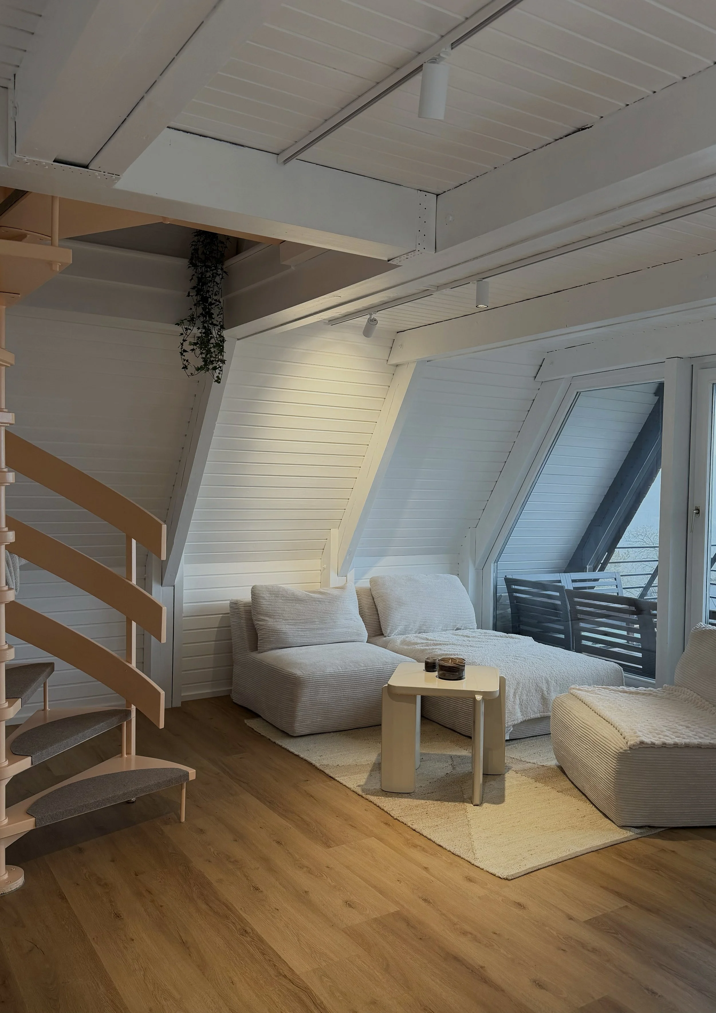 Ein gemütliches Wohnzimmer mit weißen Wänden, einem großen Fenster, einem bequemen weißen Sofa, einem kleinen weißen Beistelltisch mit drei Gläsern, einem hellen Teppich und einem Holzfußboden.