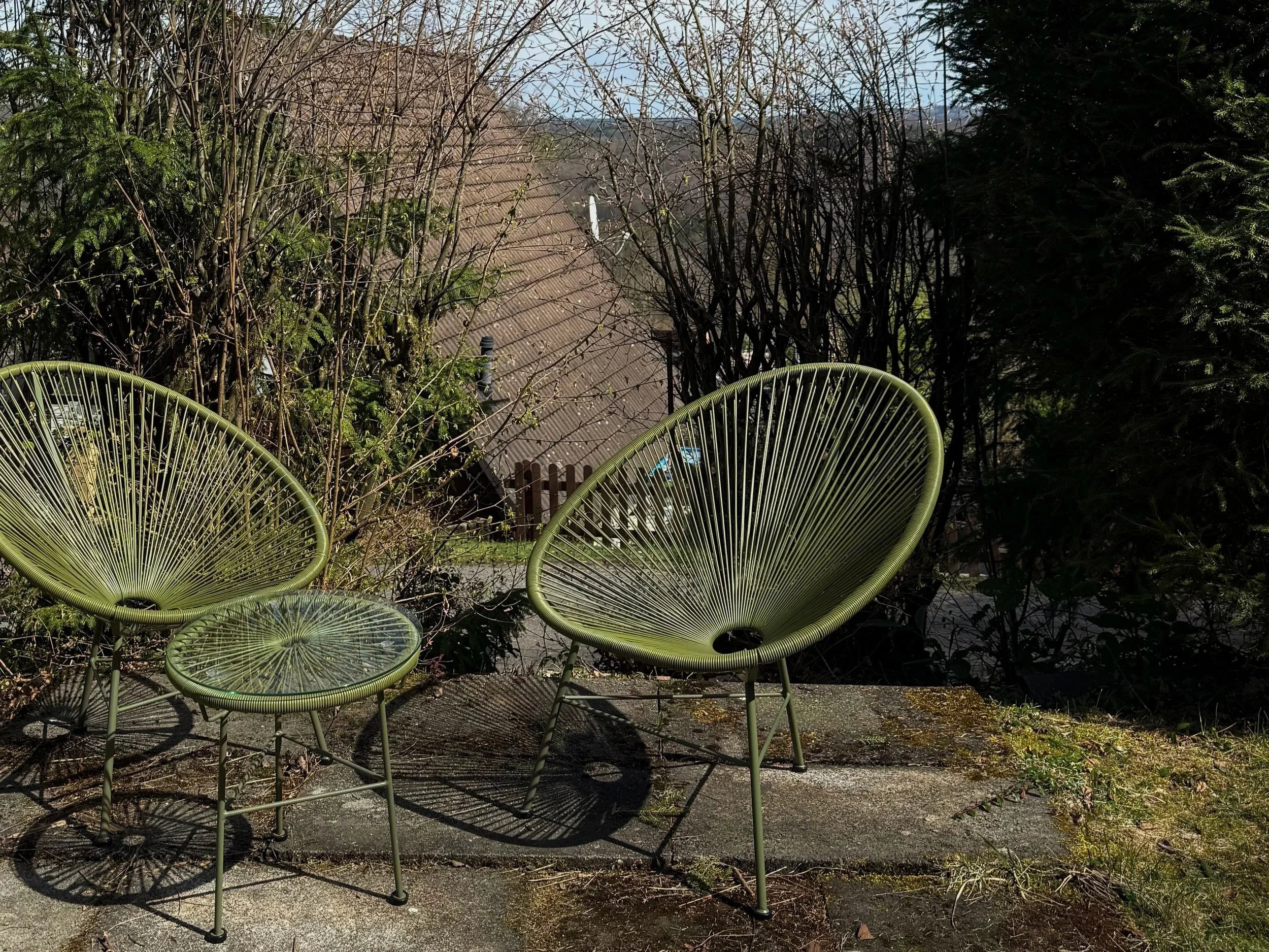 Draufsicht auf grüne Stühle im Garten mit Bäumen, Dach eines Hauses im Hintergrund, sonniges Wetter.