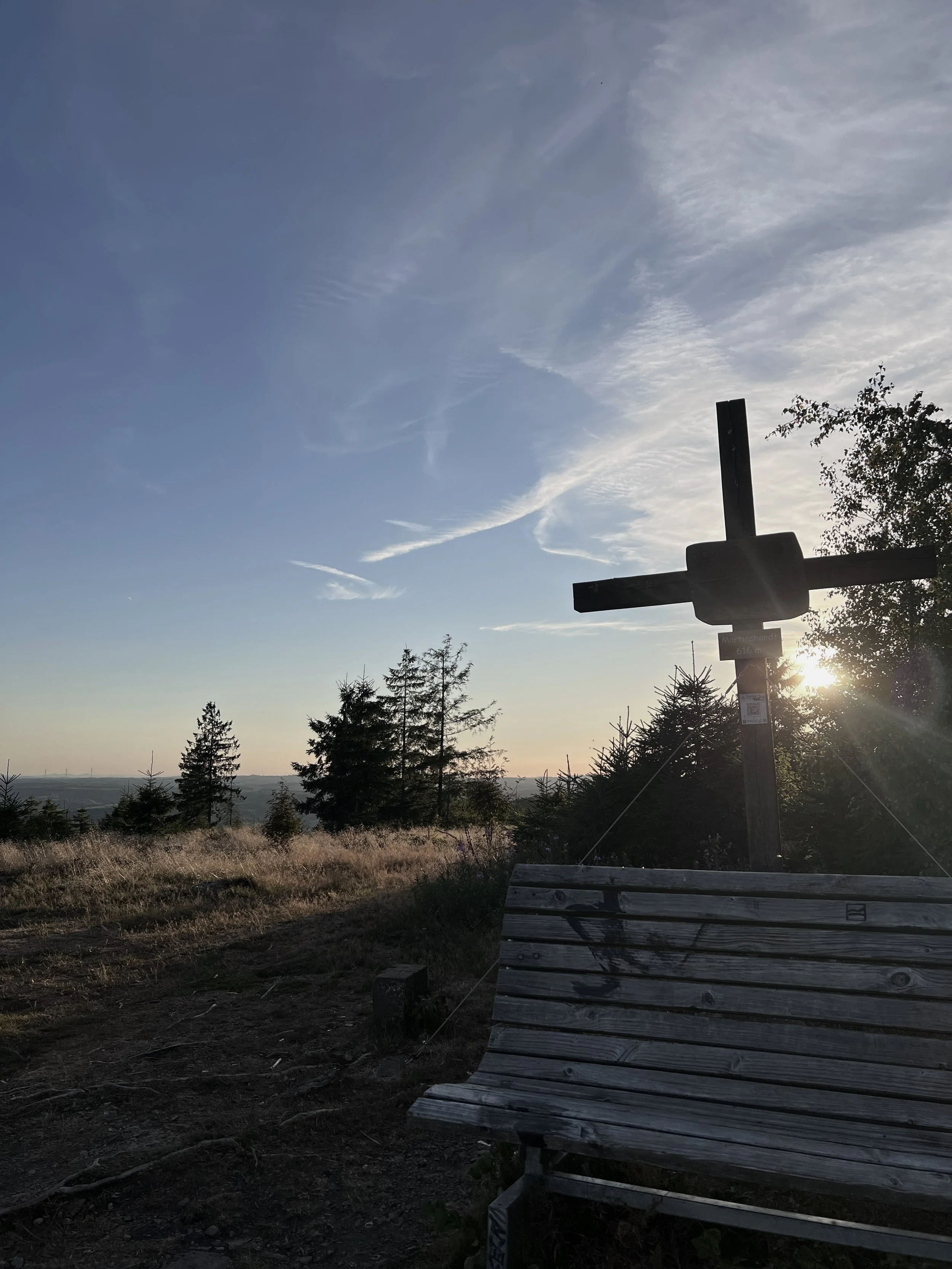 Ein Holzbank und ein Holzkreuz vor einem Sonnenuntergang bei einem Hügel mit Bäumen, blauer Himmel mit einigen Wolken.