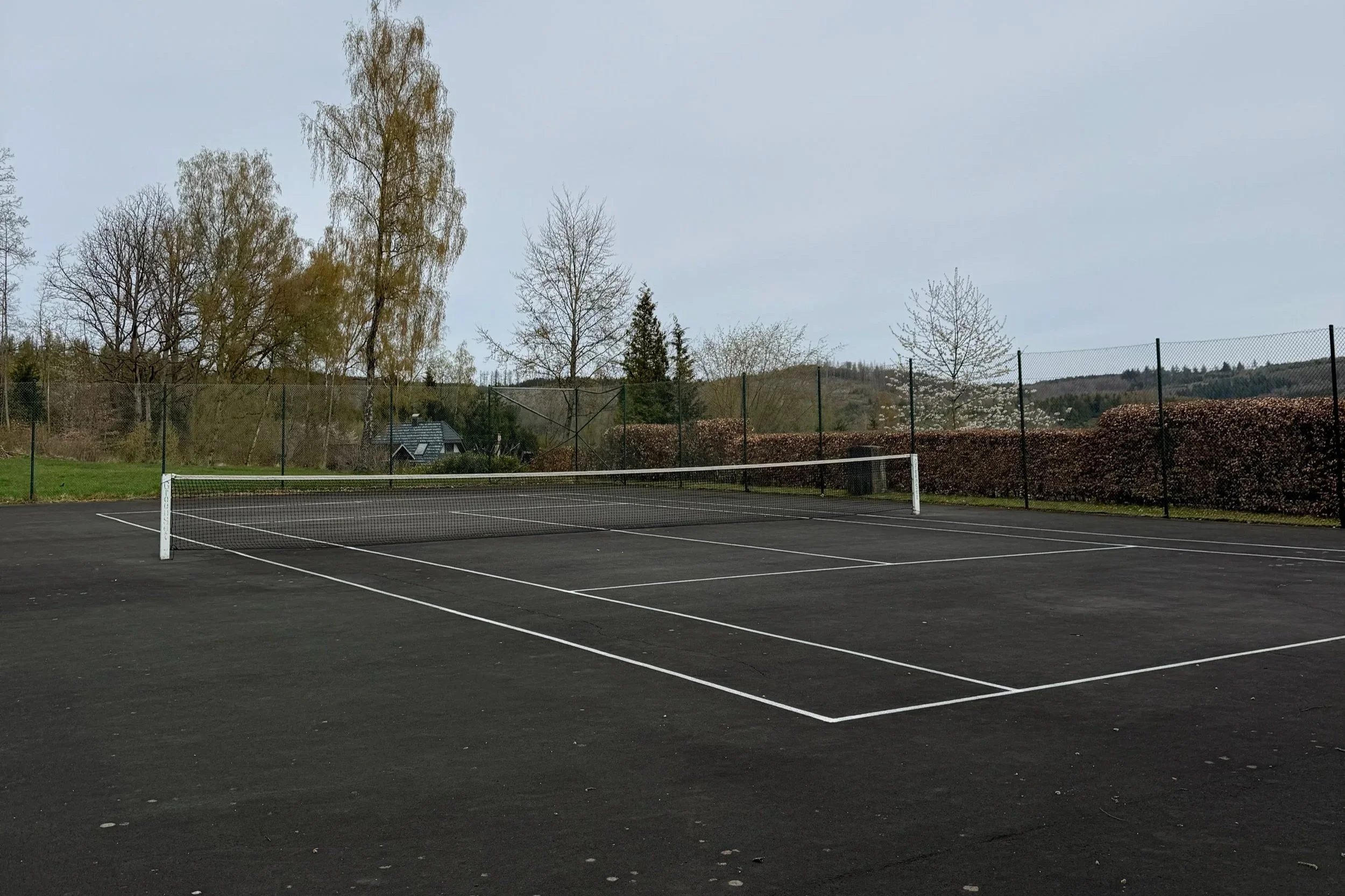 Leerer Tennisplatz im Freien mit Netzen und umgeben von Bäumen und Hügeln, bewölkter Himmel.