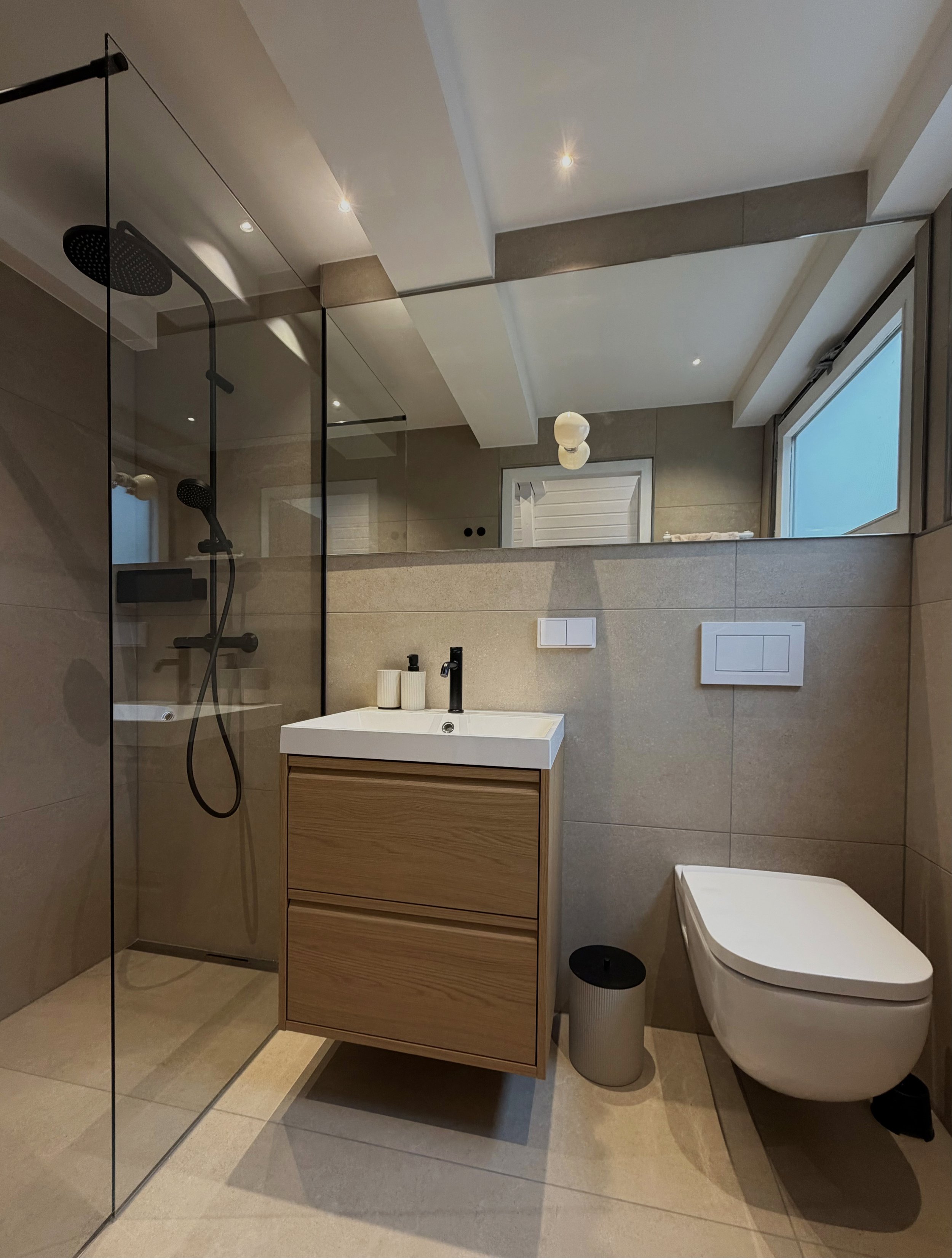 Moderner Badezimmerbereich mit Dusche, Waschtisch, Wandhocktoilette, dunkler Badmülleimer, großen Spiegeln und Fenster, das Tageslicht hereinlässt.