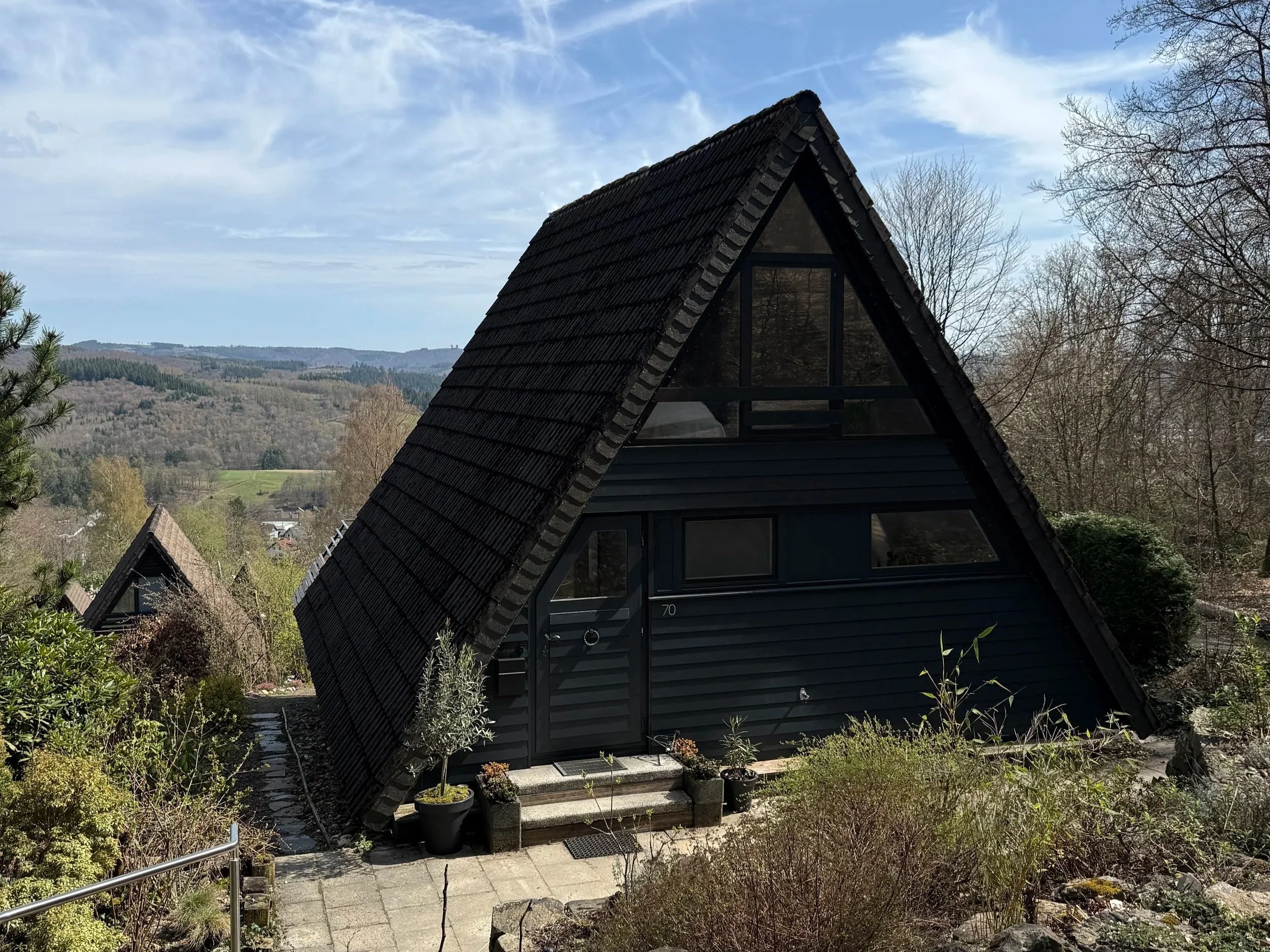Ein modernes Haus mit einem steilen, schindelverkleideten A-Raum-Dach, umgeben von Bäumen und einer Gartenlandschaft, mit Blick auf eine Hügel- und Waldlandschaft im Hintergrund.