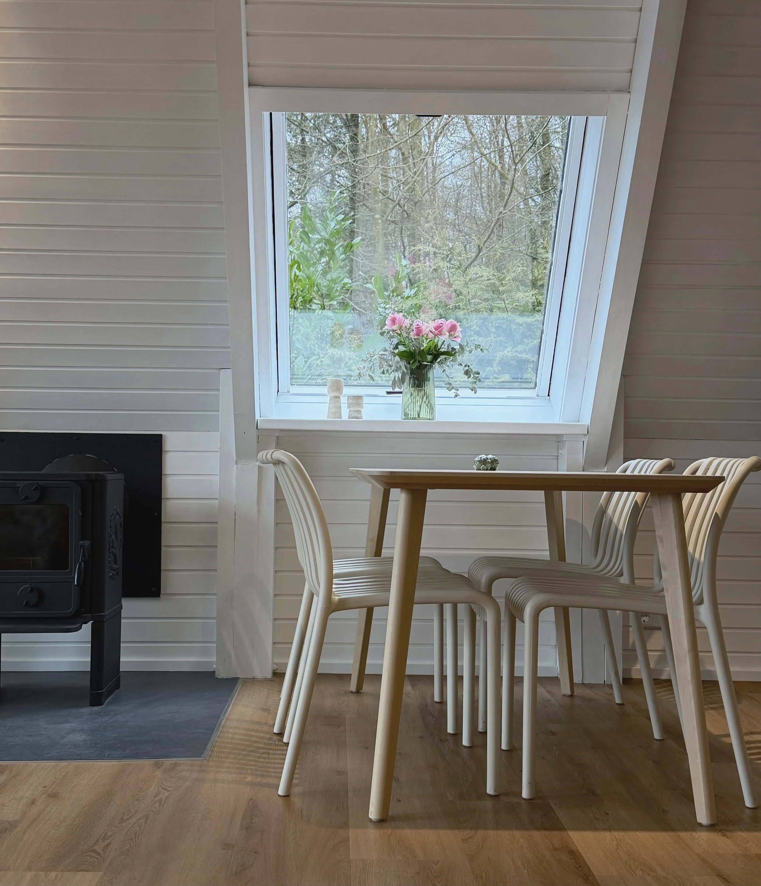 Inneneinrichtung mit Esstisch, weißen Stühlen und Fenster mit Blick auf Bäume und blühende Pflanzen im Hintergrund.