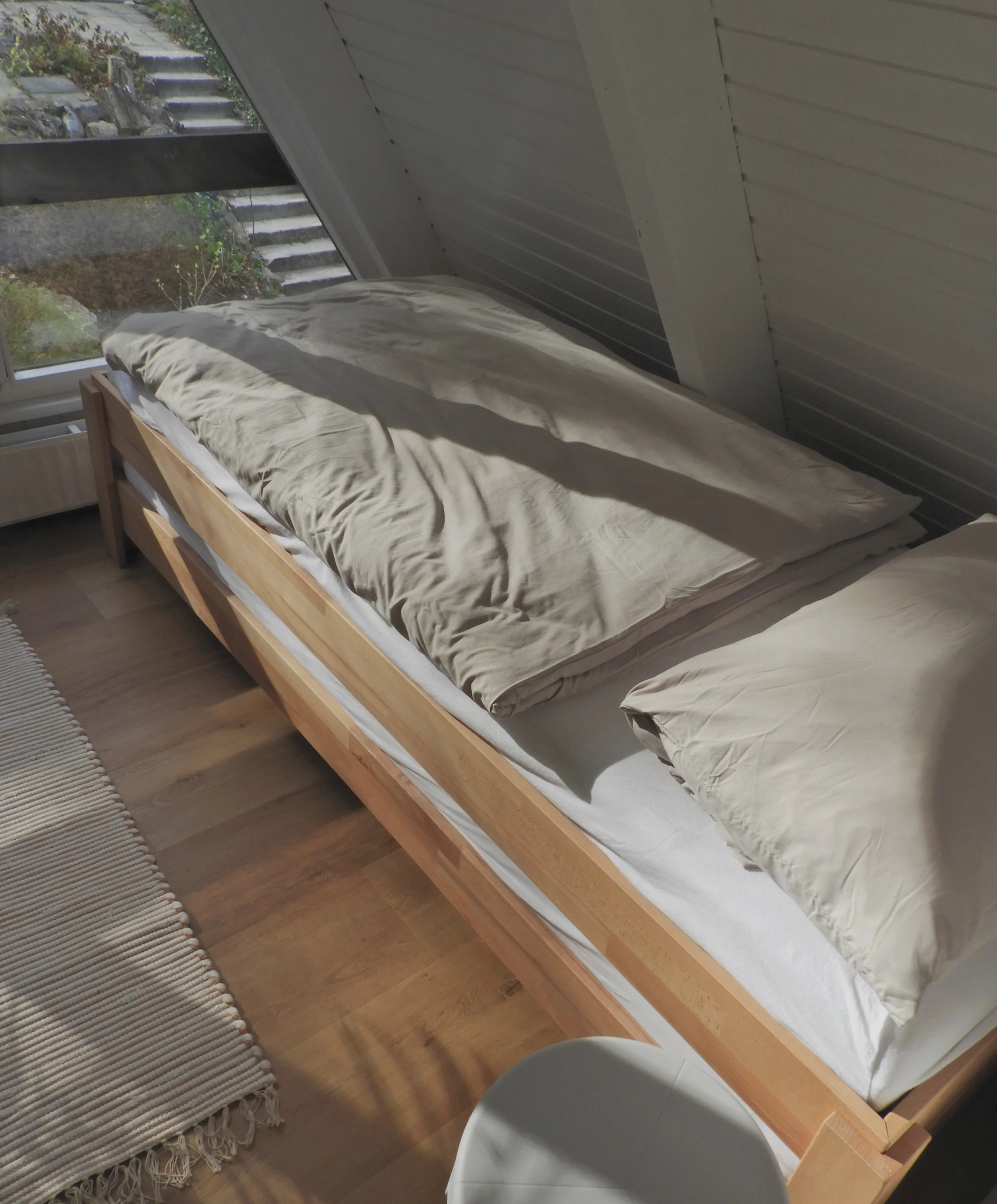Ein Schlafzimmer mit einem Holzrahmenbett, beige Bettwäsche, einem großen Fenster mit Blick auf Steinstufen und Pflanzen im Außenbereich, einem Teppich neben dem Bett und einem weißen Stuhl im Vordergrund.