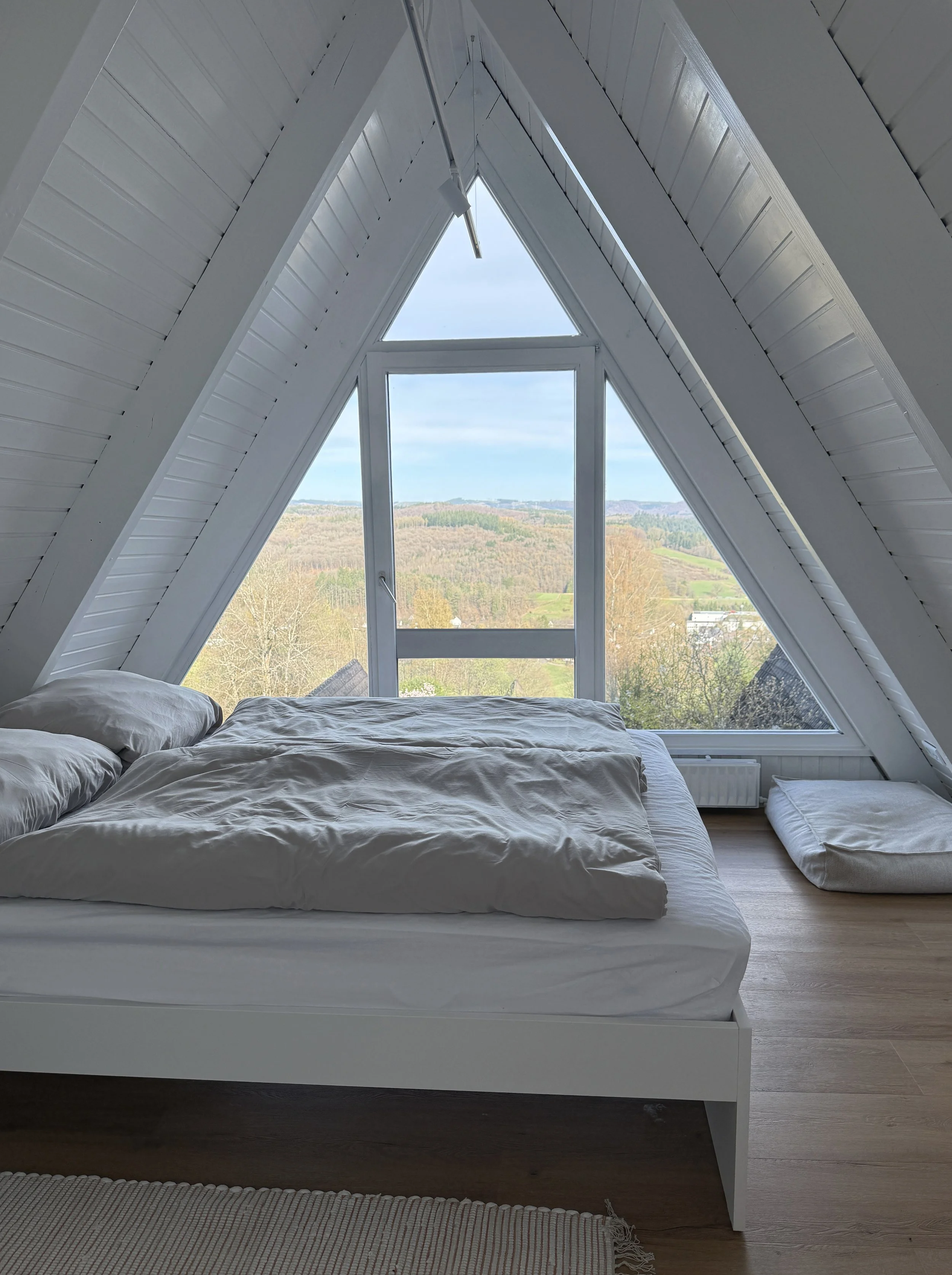 Ein Schlafzimmer mit weißem Bett, das vor einem großen, dreieckigen Fenster liegt. Im Freien sieht man eine weite Landschaft mit Bäumen und Hügeln, es ist bewölkt.