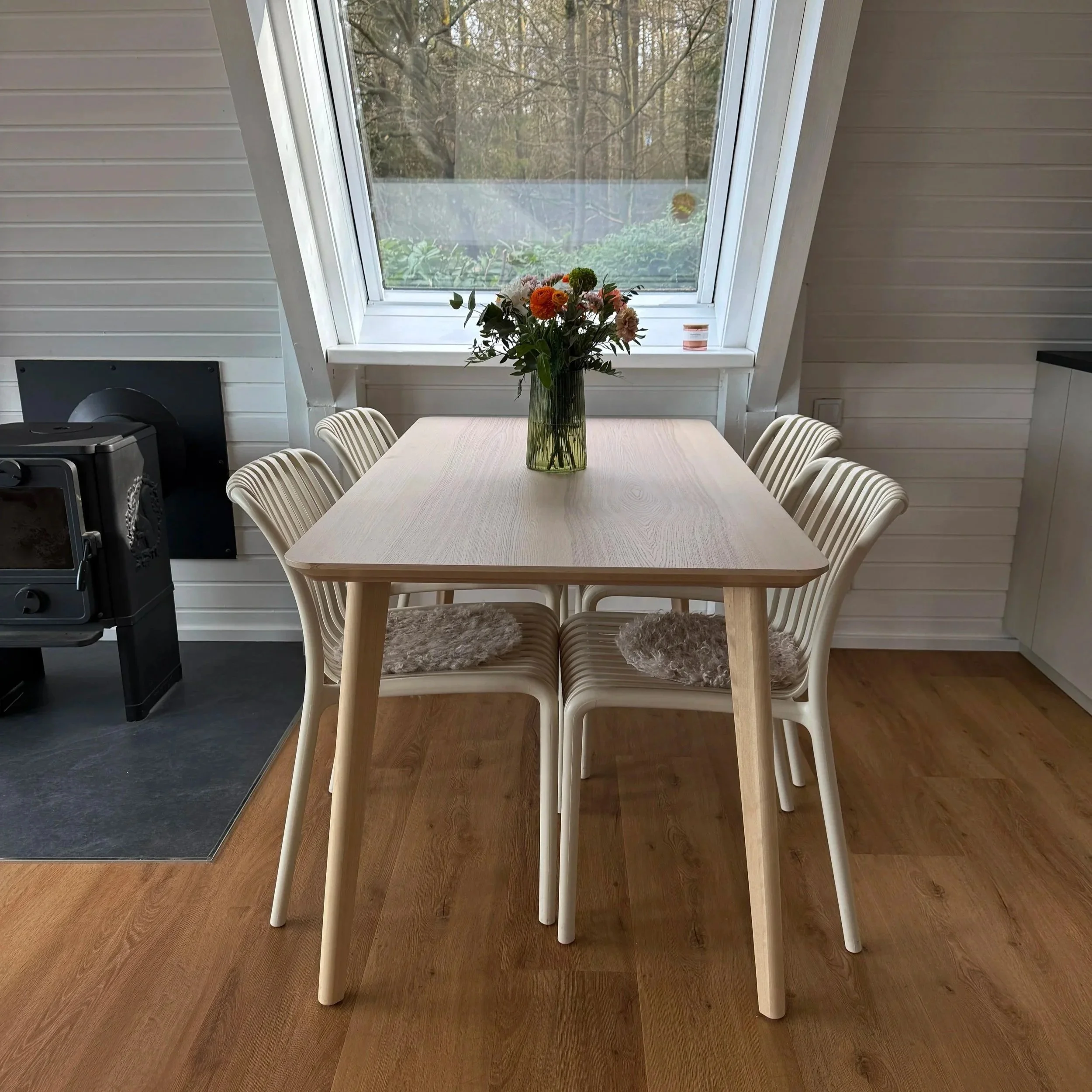 Ein Esstisch mit weißen Stühlen, einer Blumenvase mit bunten Blumen und einer Fensterscheibe mit Blick nach draußen auf Bäume und Himmel.