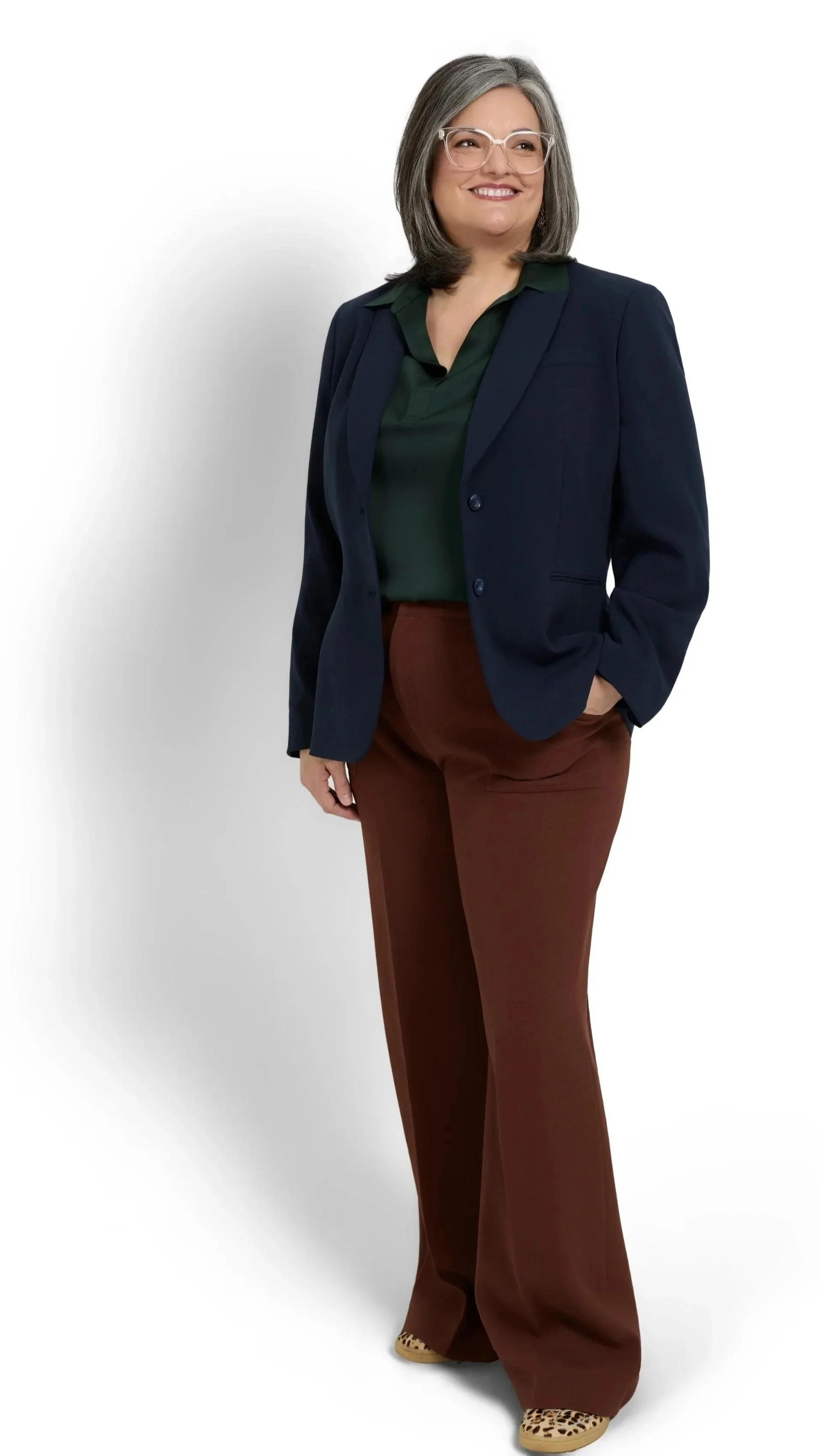 A woman with gray hair, glasses, wearing a navy blazer, green blouse, brown wide-leg pants, and leopard print shoes, smiling and standing with one hand in her pocket against a white background.