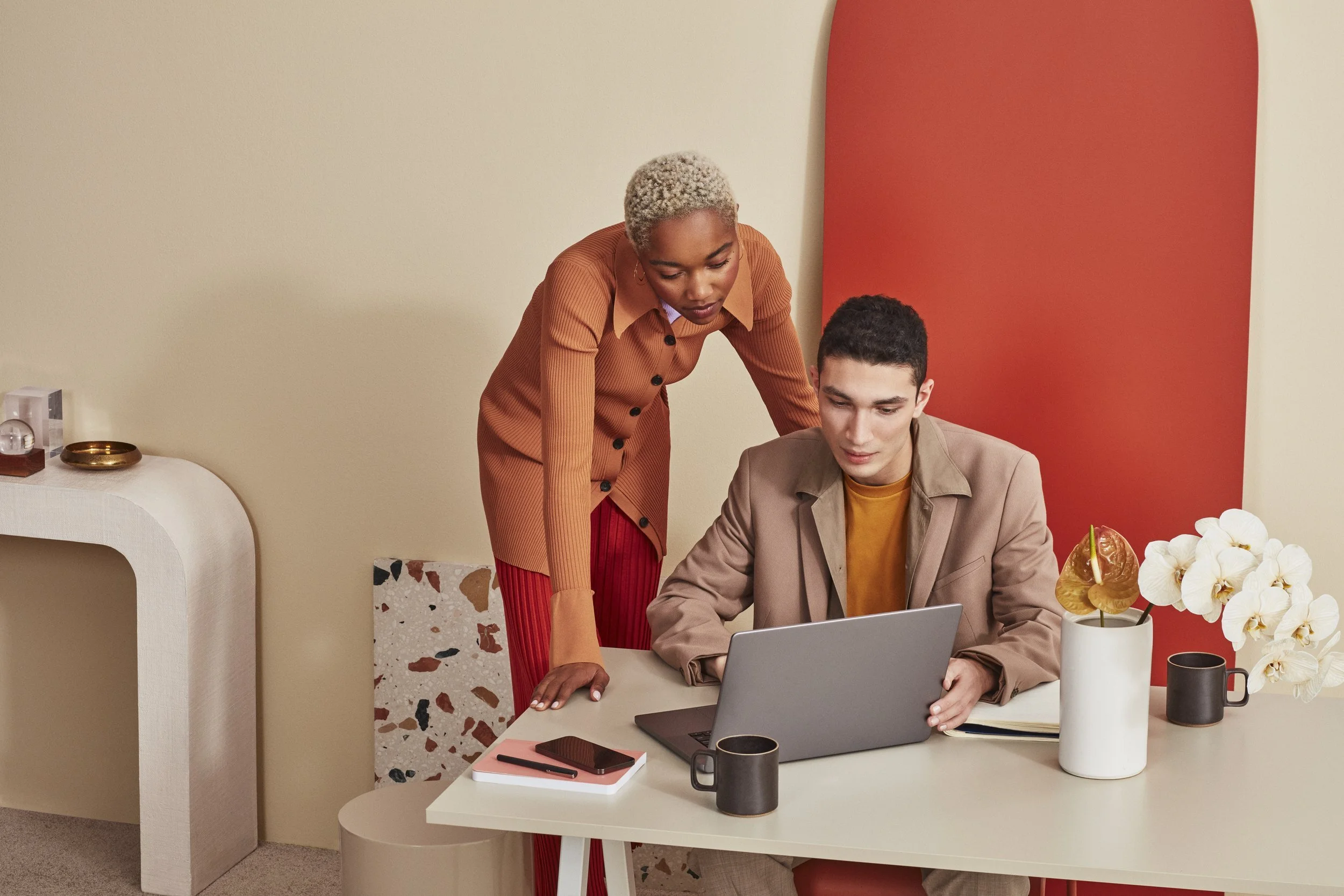 Two people working together at a desk using a laptop, with coffee mugs, a notepad, and flowers on the table, in a modern office setting.
