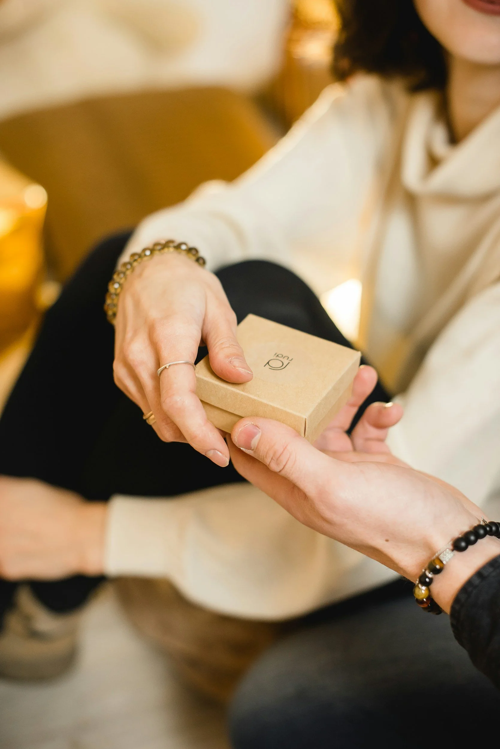 Two people exchanging a small tan box with a black logo, possibly during a gift exchange or special occasion.