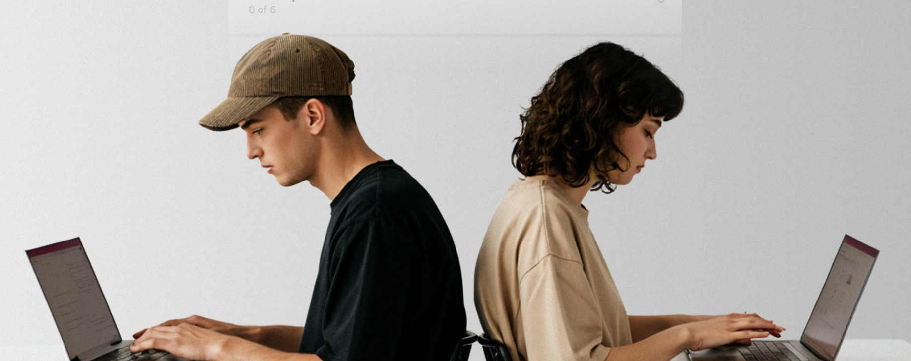 A young man and woman sitting back-to-back, each working on a laptop against a plain light gray background.
