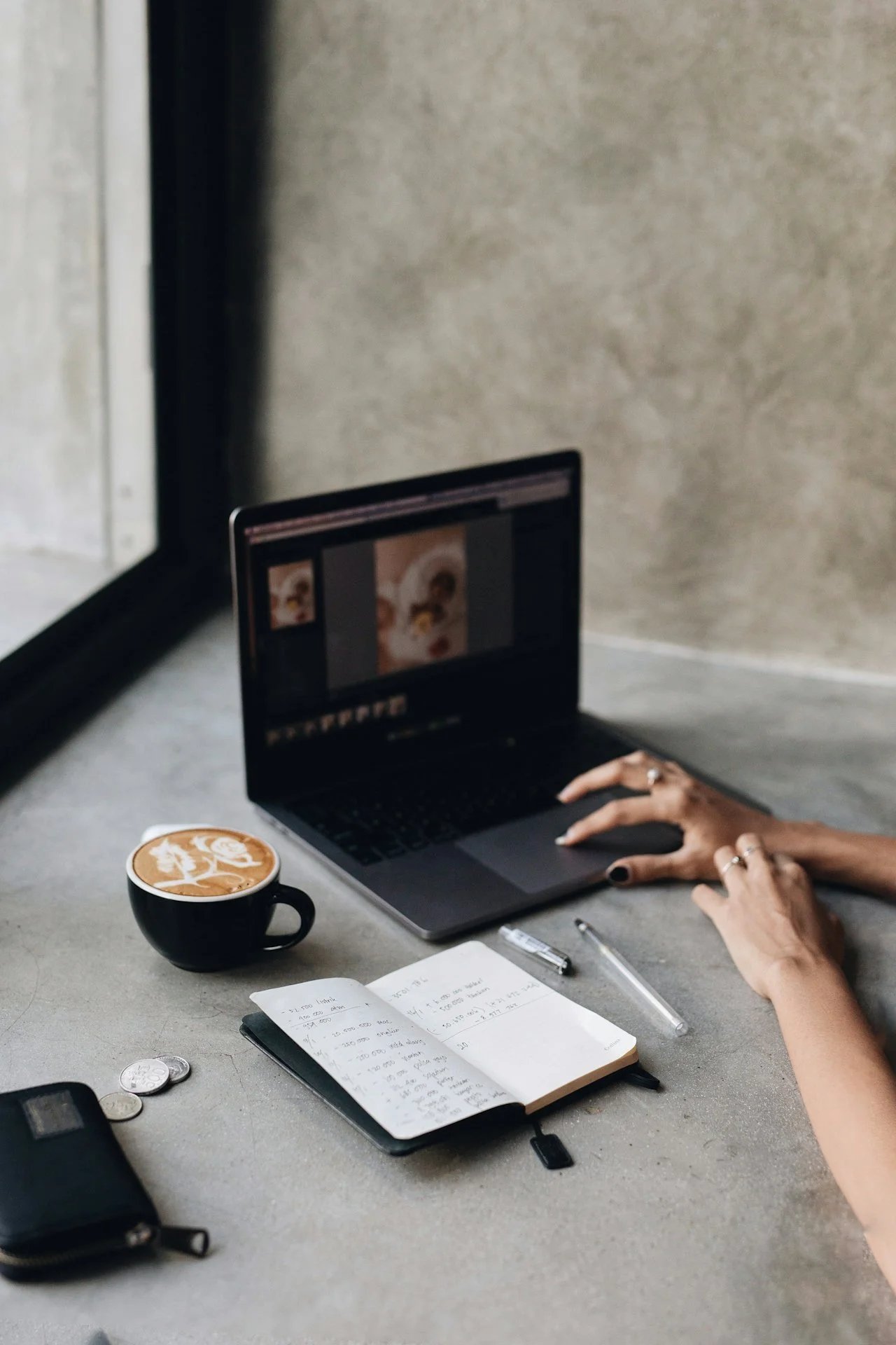 Close-up of ringed hands typing on a laptop at a concrete desk alongside a latte and open notebook, representing the focused custom website build phase of Knight Theory™'s Full Court brand and Squarespace web design package.